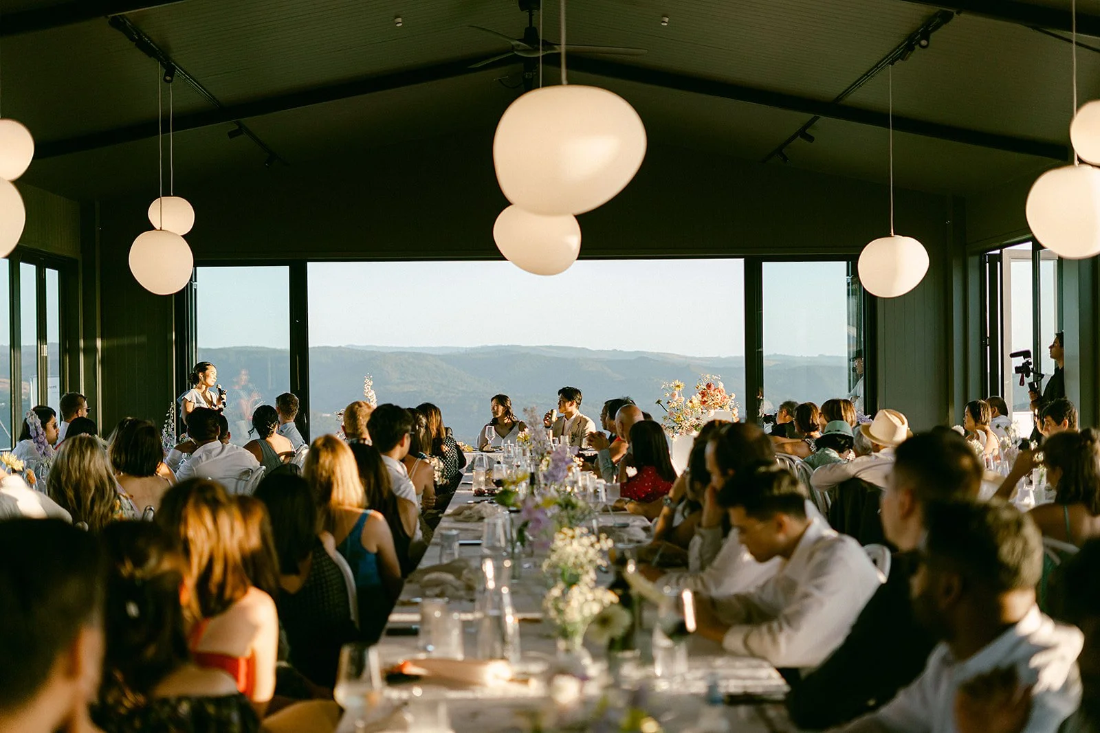 Guests gather for a formal dinner and speech in a glass-walled venue overlooking forested mountains.