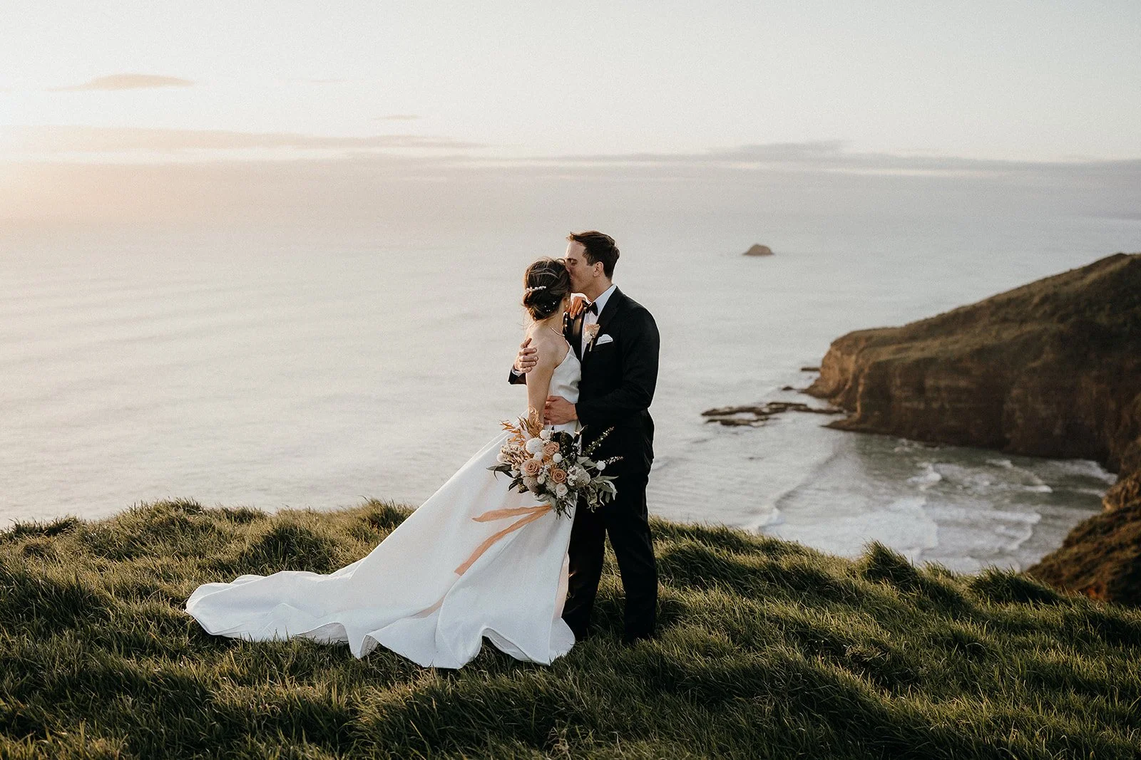 Newlywed couple embracing on a grassy coastal cliff overlooking the ocean