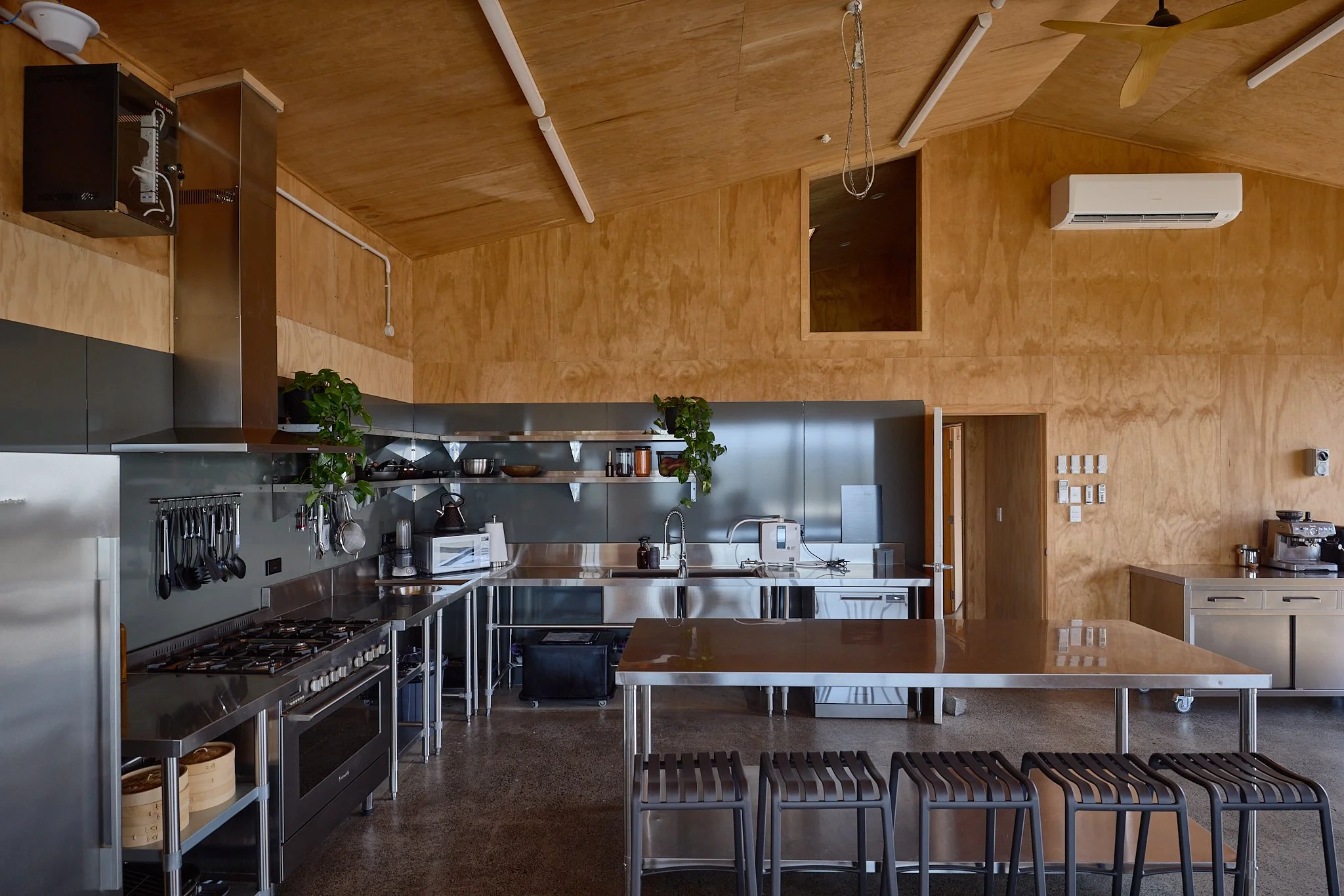 Modern commercial kitchen with stainless steel appliances, large island, cookware, bar stools, and wooden walls