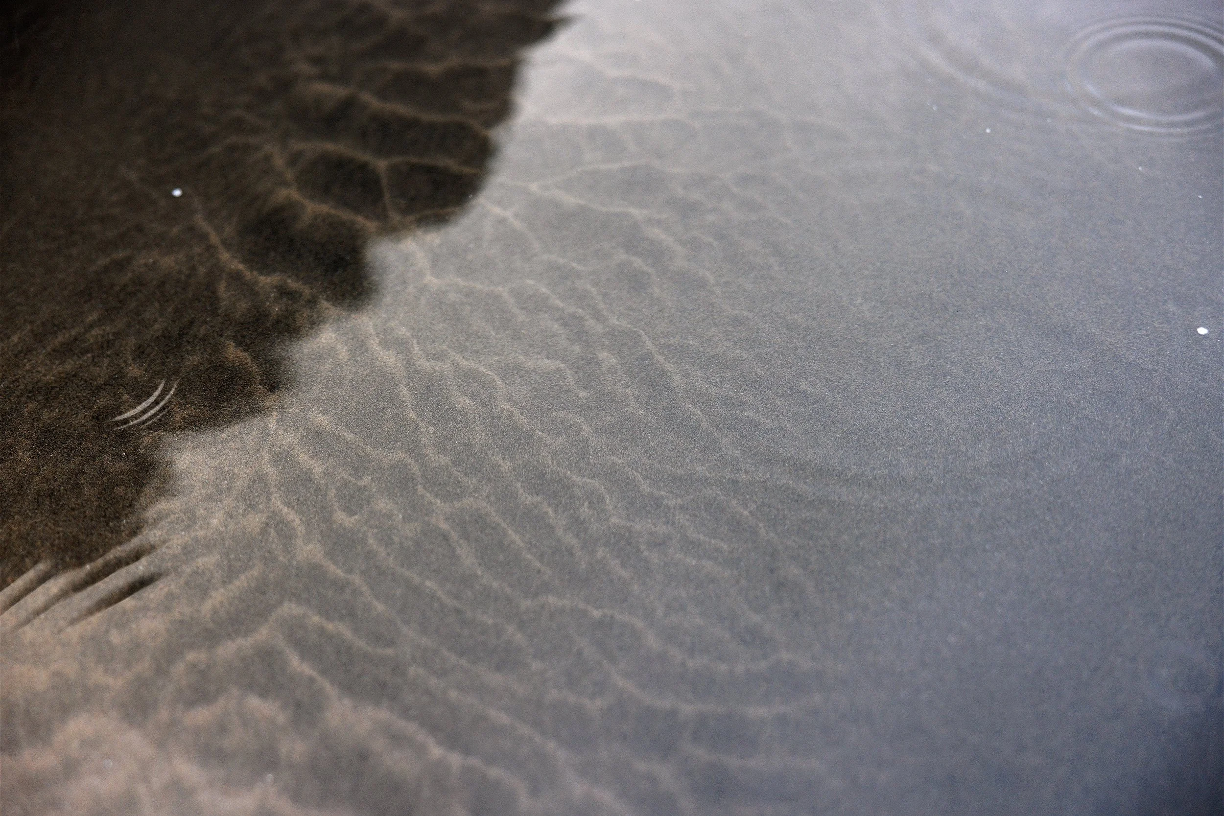 Close-up of dark sand meeting shallow rippling water on a beach