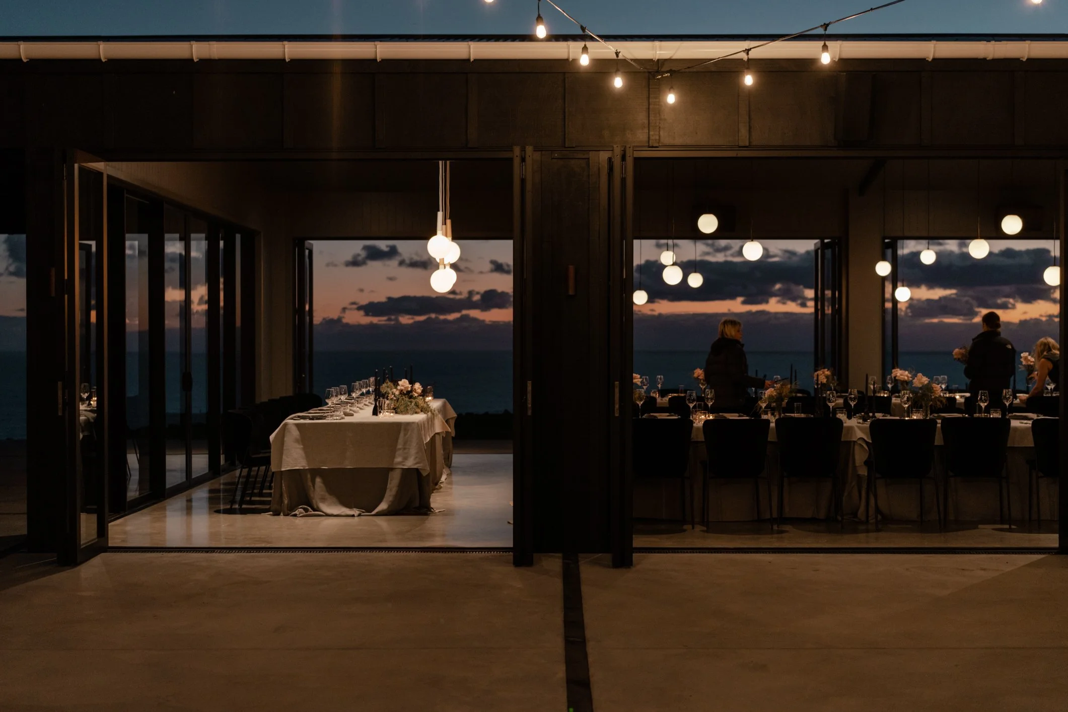 Elegant oceanfront dining room with candlelit tables, floral centerpieces, pendant lights, and sunset views