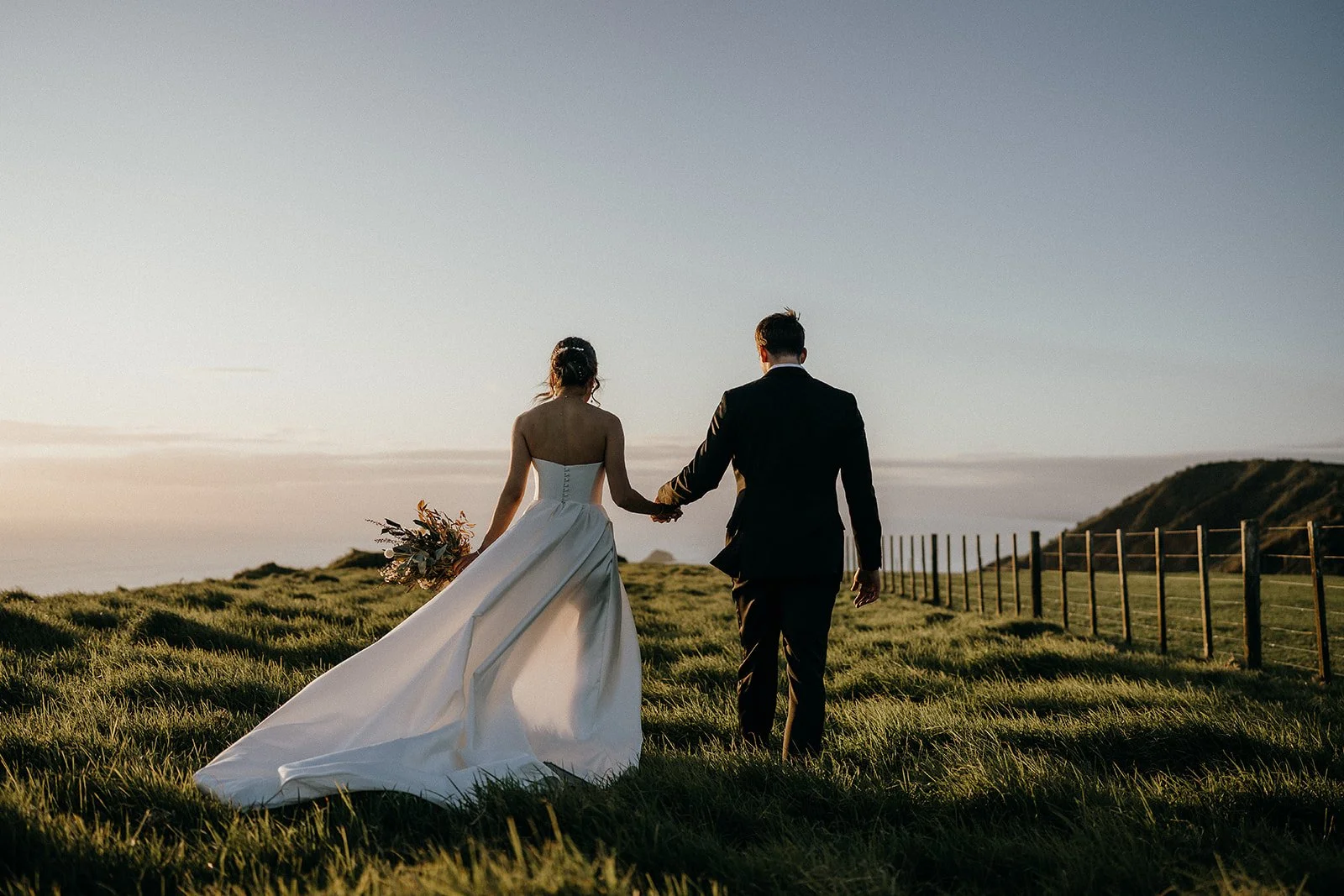 Newlywed couple holding hands and walking through a grassy coastal field at sunset, bride carrying a bouquet.