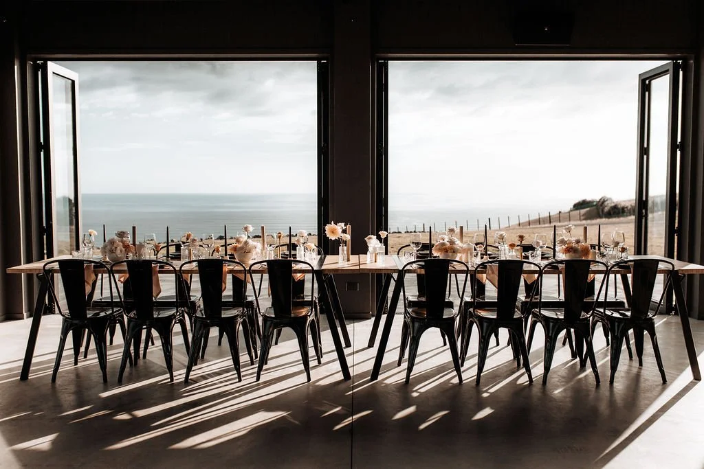 Elegant seaside restaurant dining room with a long decorated table, black chairs, open windows, and ocean views