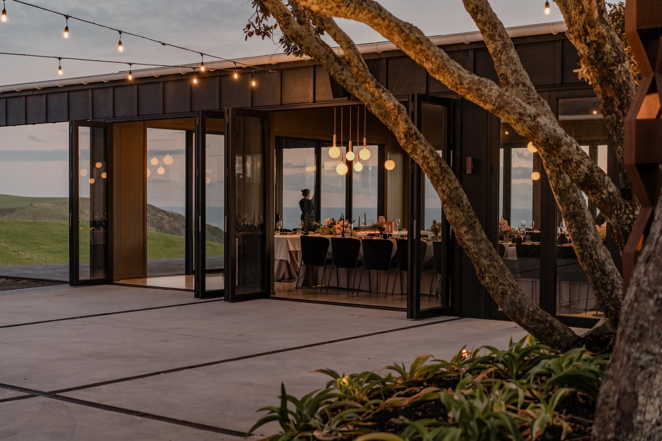 Modern glass-walled restaurant overlooking coastal hills at dusk, with string lights, pendant lamps, and outdoor dining tables.