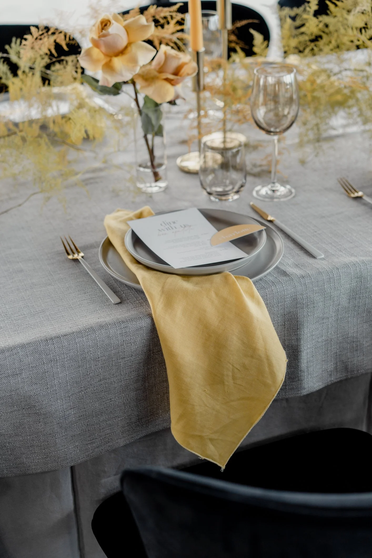 Elegant wedding table setting with yellow napkin, gray plates, menu card, glassware, peach roses, and floral greenery