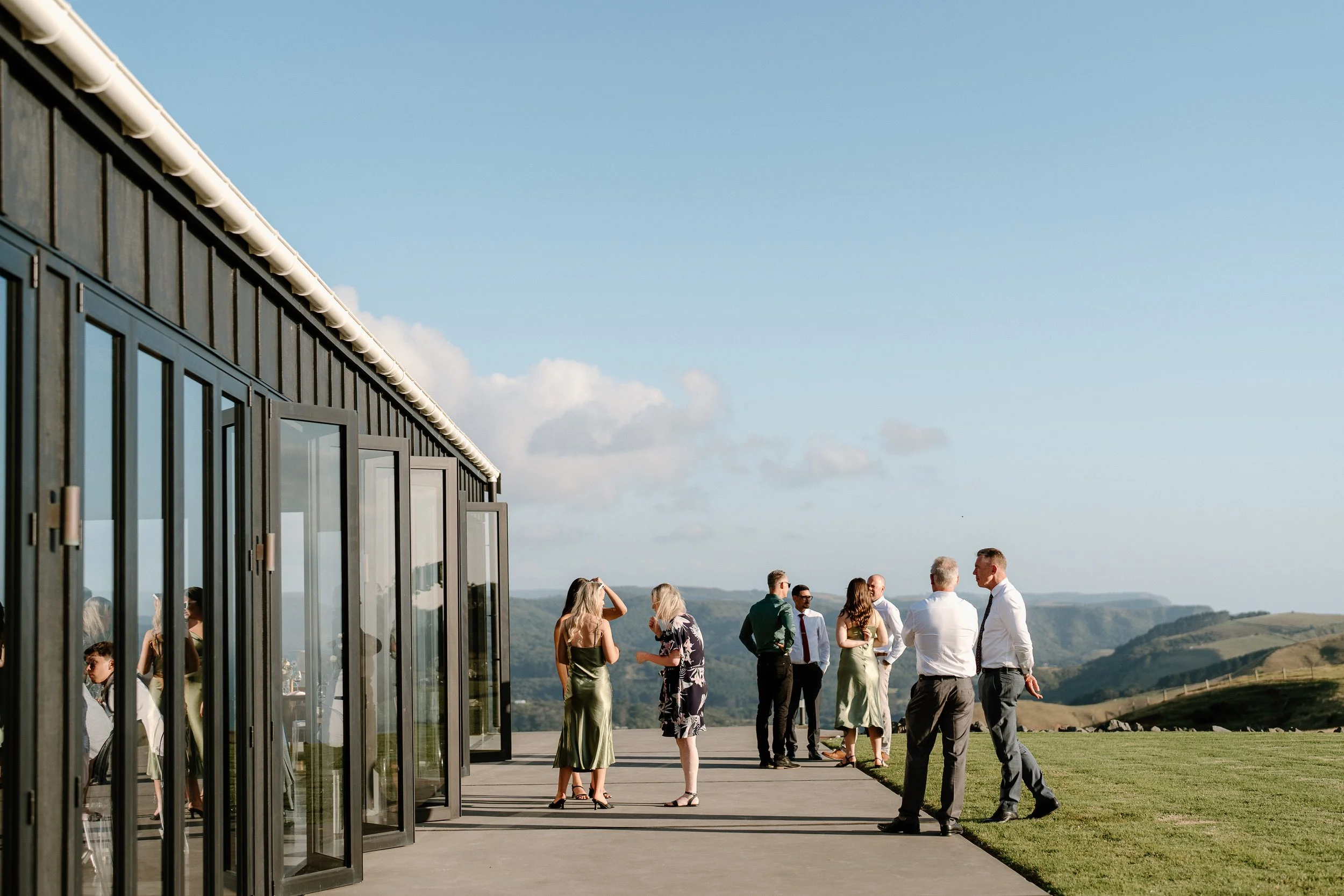 Wedding guests gather outside a modern glass building overlooking rolling hills.