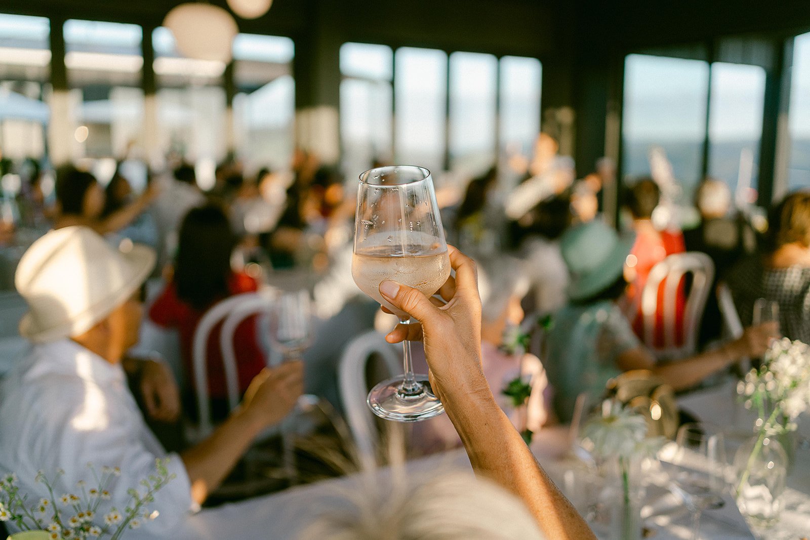 Hand raising a stemmed glass of white wine at a sunlit outdoor gathering