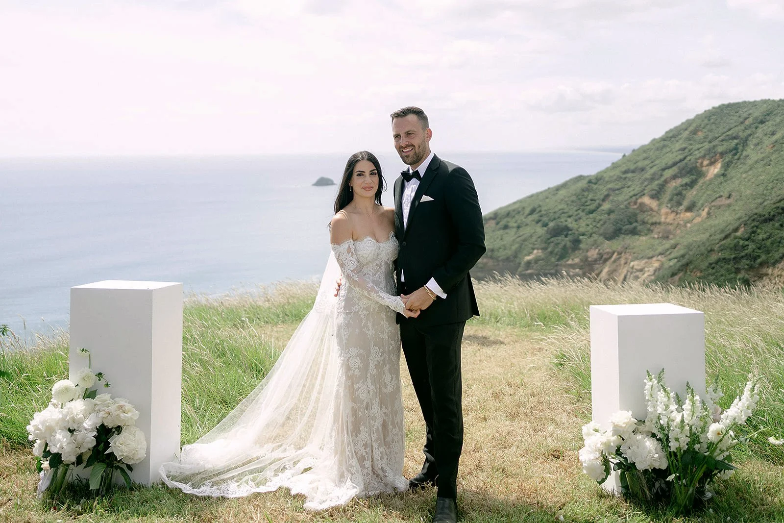 Newlywed couple posing in wedding attire at a seaside cliff wedding ceremony with white floral arrangements.