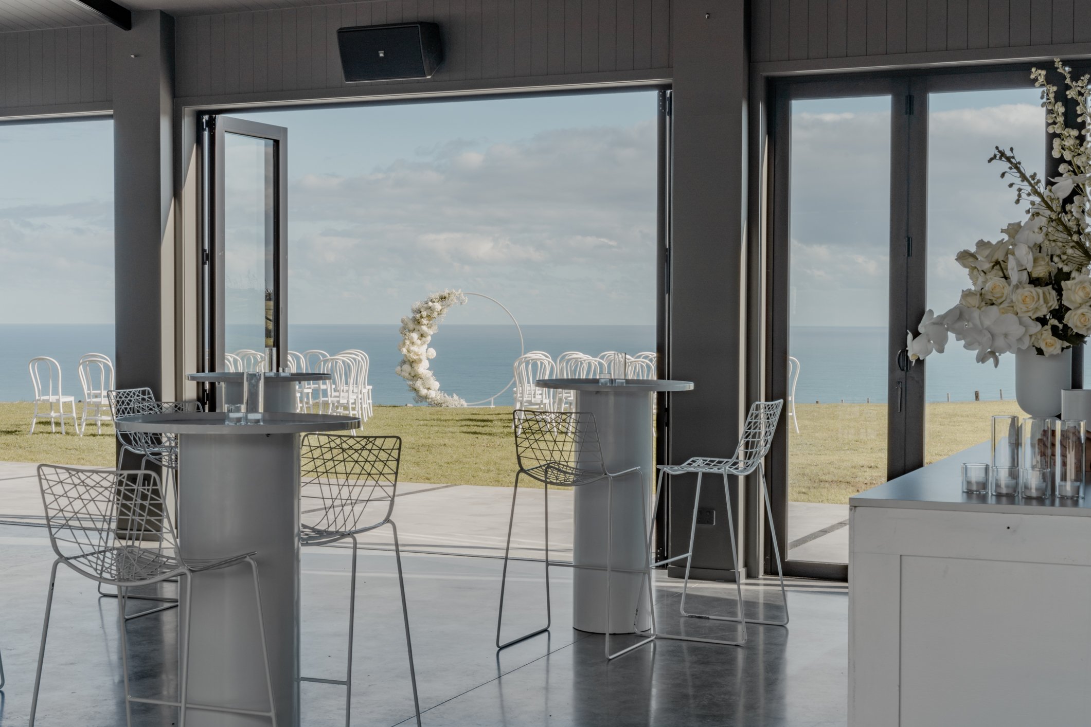 Modern seaside wedding venue with cocktail tables, wire chairs, floral arch, and ocean views