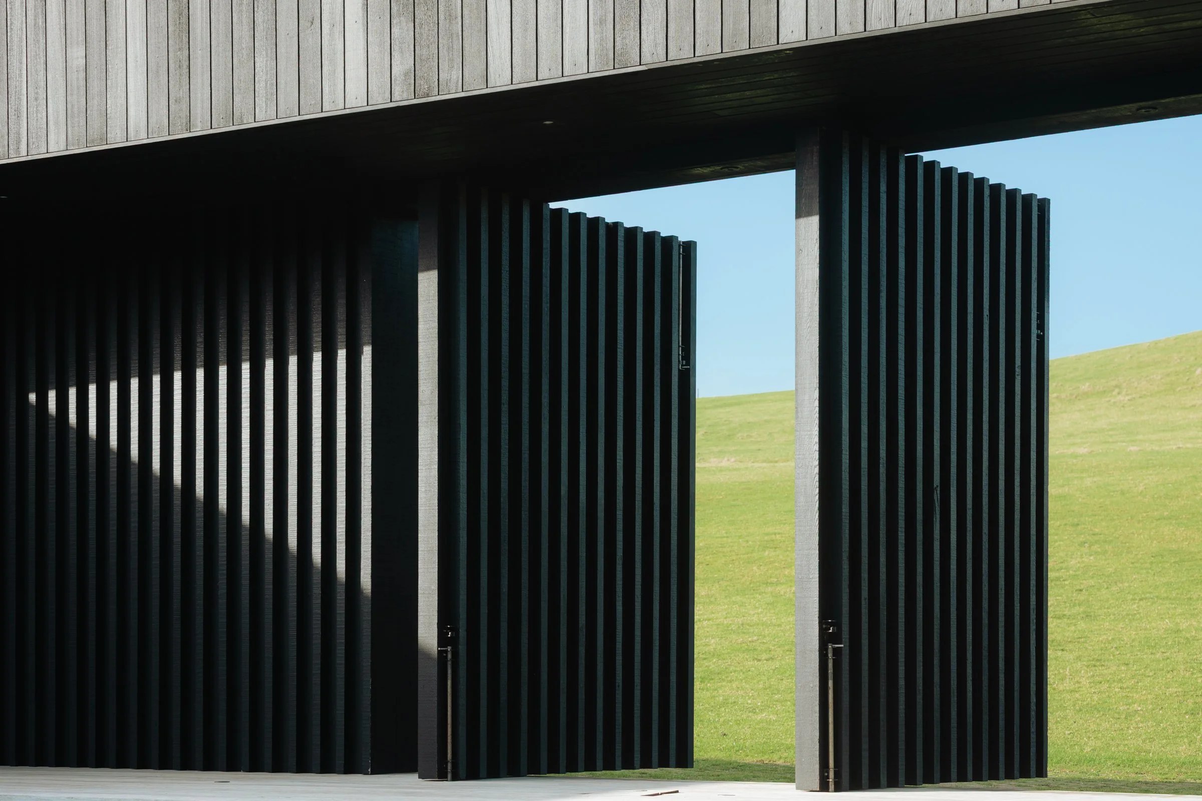 Modern black ribbed architectural structure with open panels, overlooking a grassy hillside under blue sky