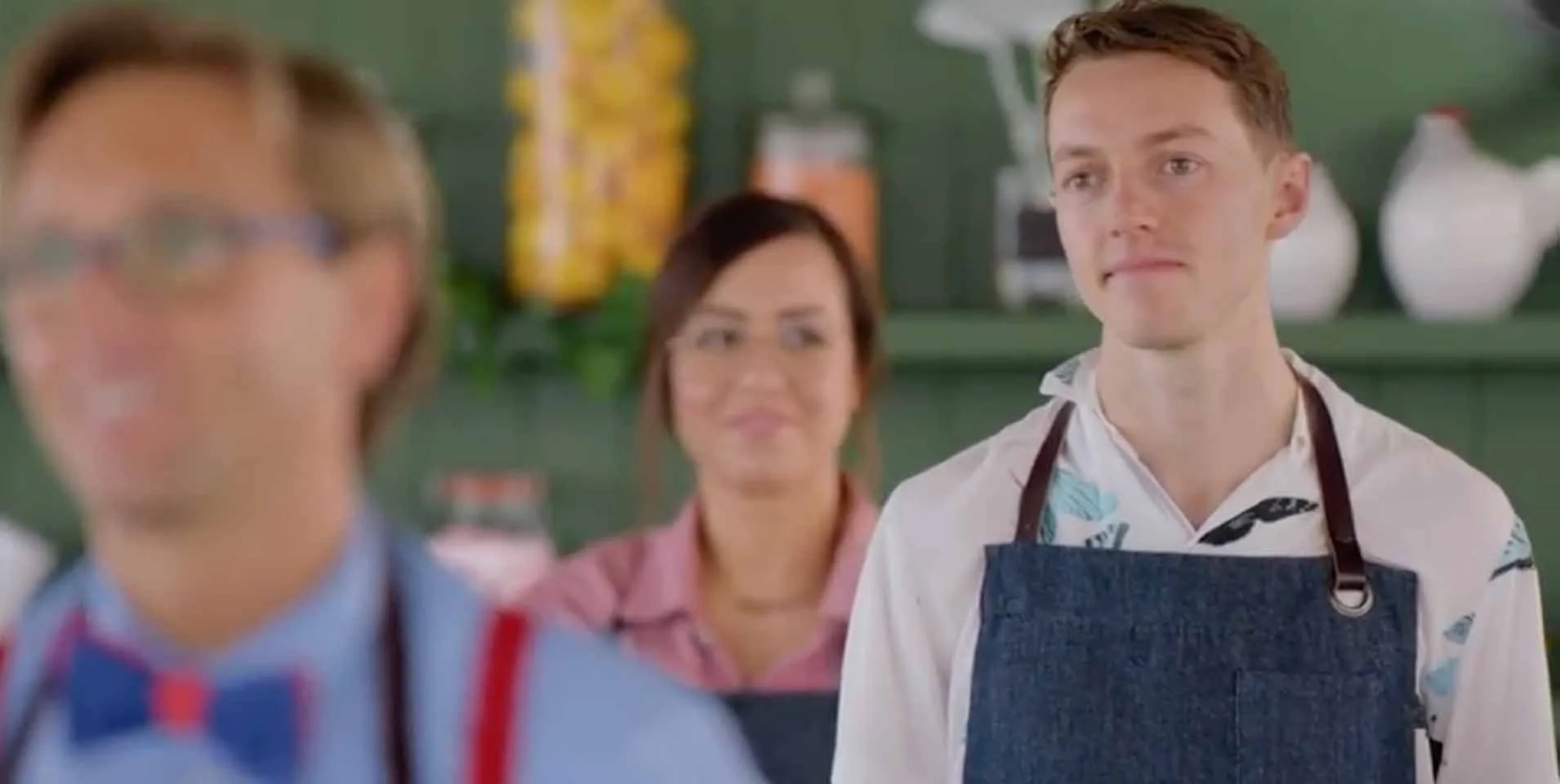 Three people in aprons standing in a kitchen, with a man in sharp focus on the right and two blurred people in the foreground and background.