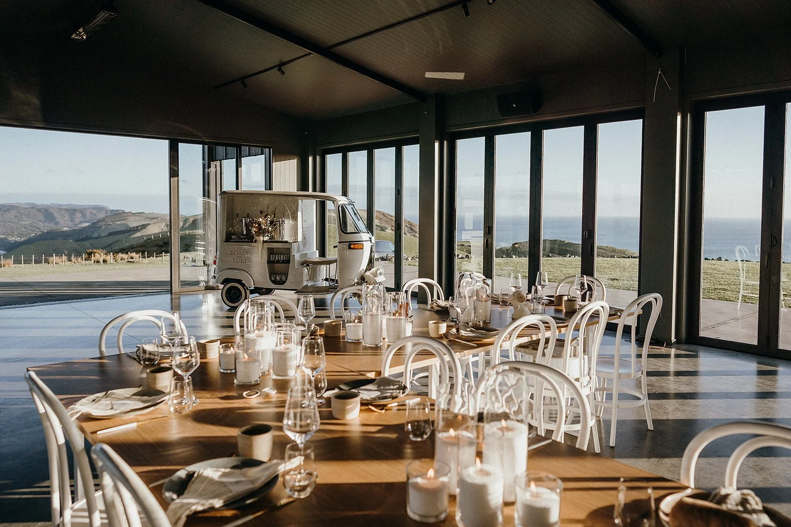 Sunlit seaside restaurant with set dining tables, a vintage food truck bar, and panoramic ocean and mountain views