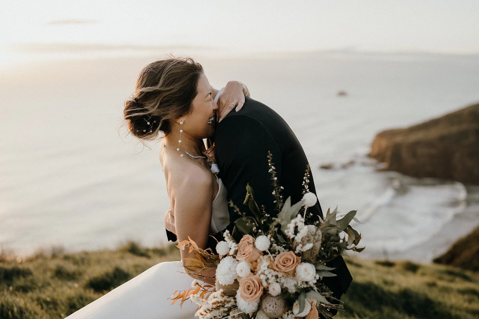 Bride and groom embracing beside the ocean, with the bride holding a bouquet of blush roses and white flowers.