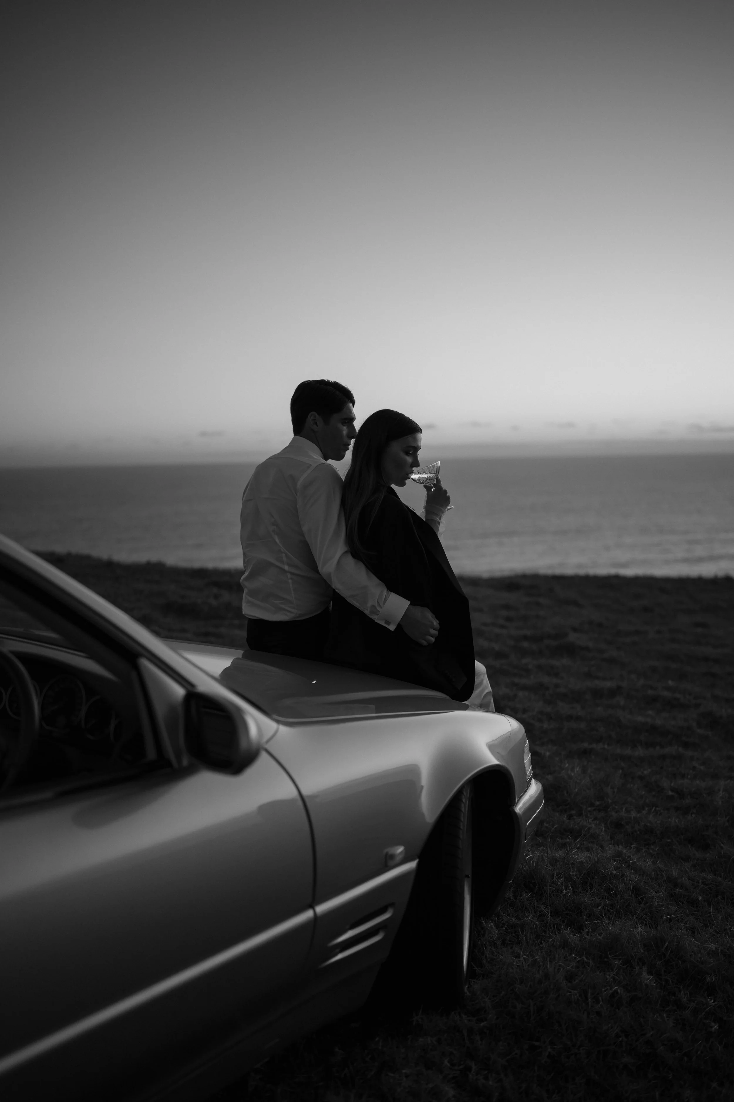 Black-and-white photo of a couple beside a sports car overlooking the ocean, with the woman drinking from a cocktail glass.