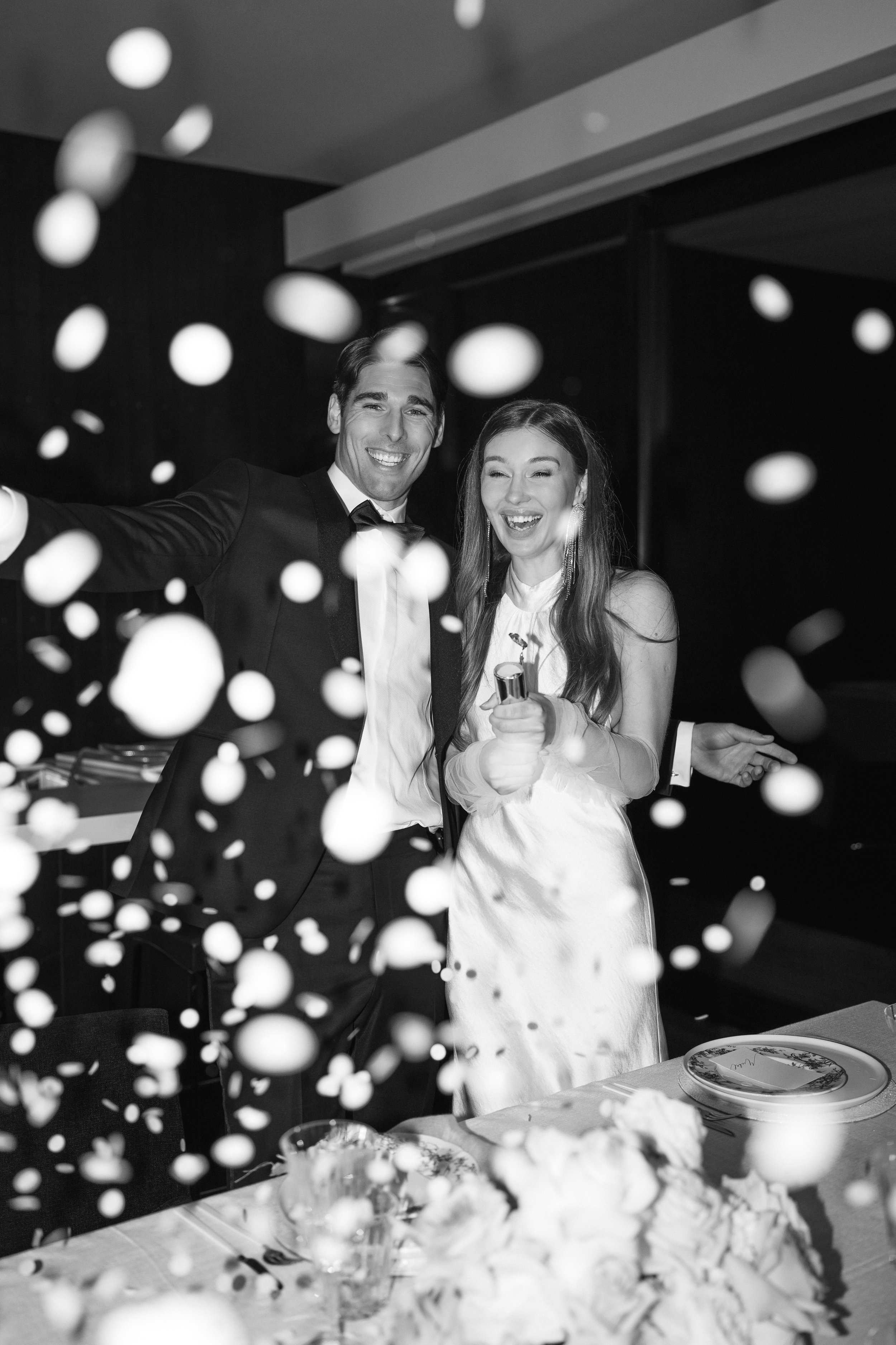 Black-and-white photo of a smiling man in a tuxedo and woman in a white dress at a dinner table, surrounded by falling confetti.