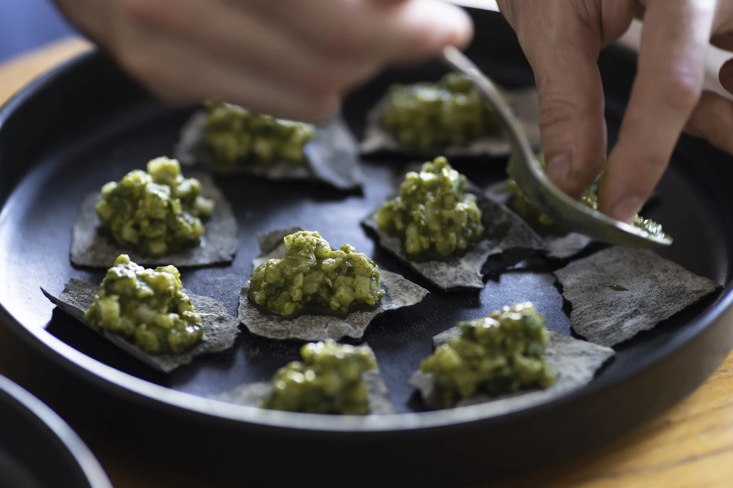 Hands spoon green avocado mixture onto black tortilla chips on a serving platter.