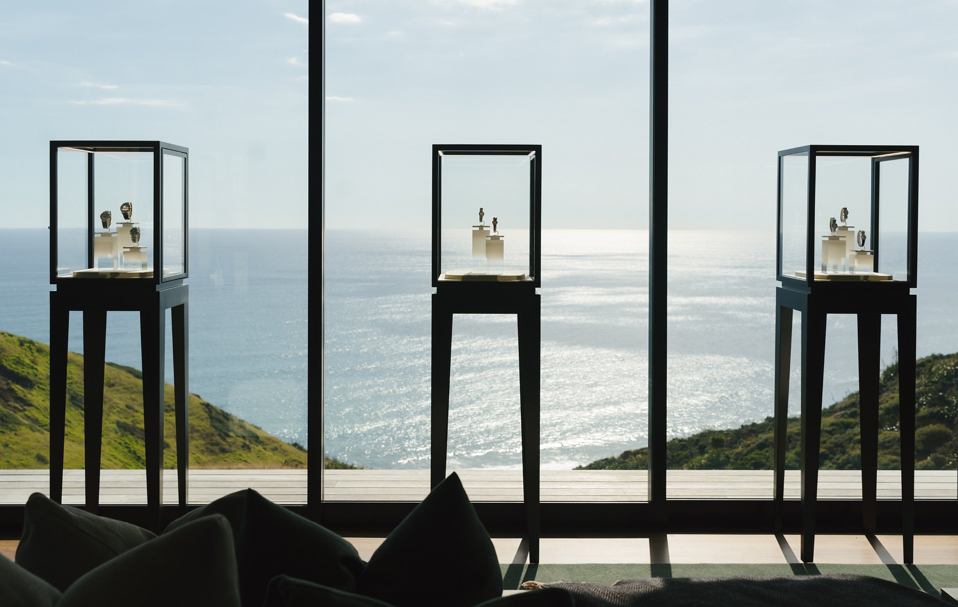 Three glass display cases holding Cartier watches in front of floor-to-ceiling windows overlooking a sunlit ocean and green coastal hills