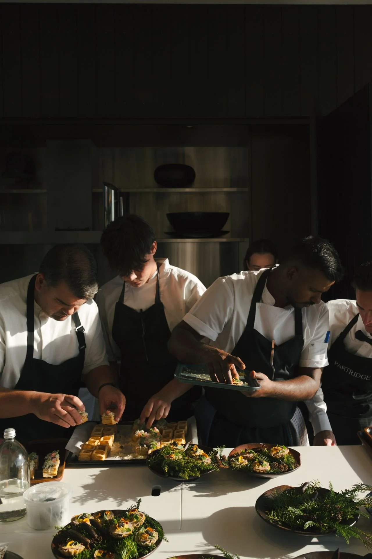 Chefs in black aprons preparing plated appetizers at a restaurant kitchen table