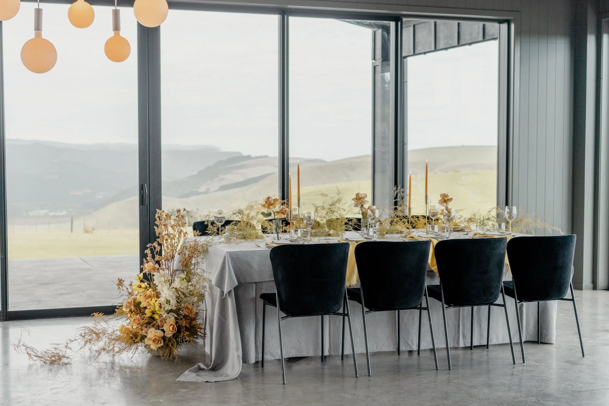Elegant wedding reception table with floral arrangements, candles, glassware, and black chairs overlooking rolling hills through floor-to-ceiling windows
