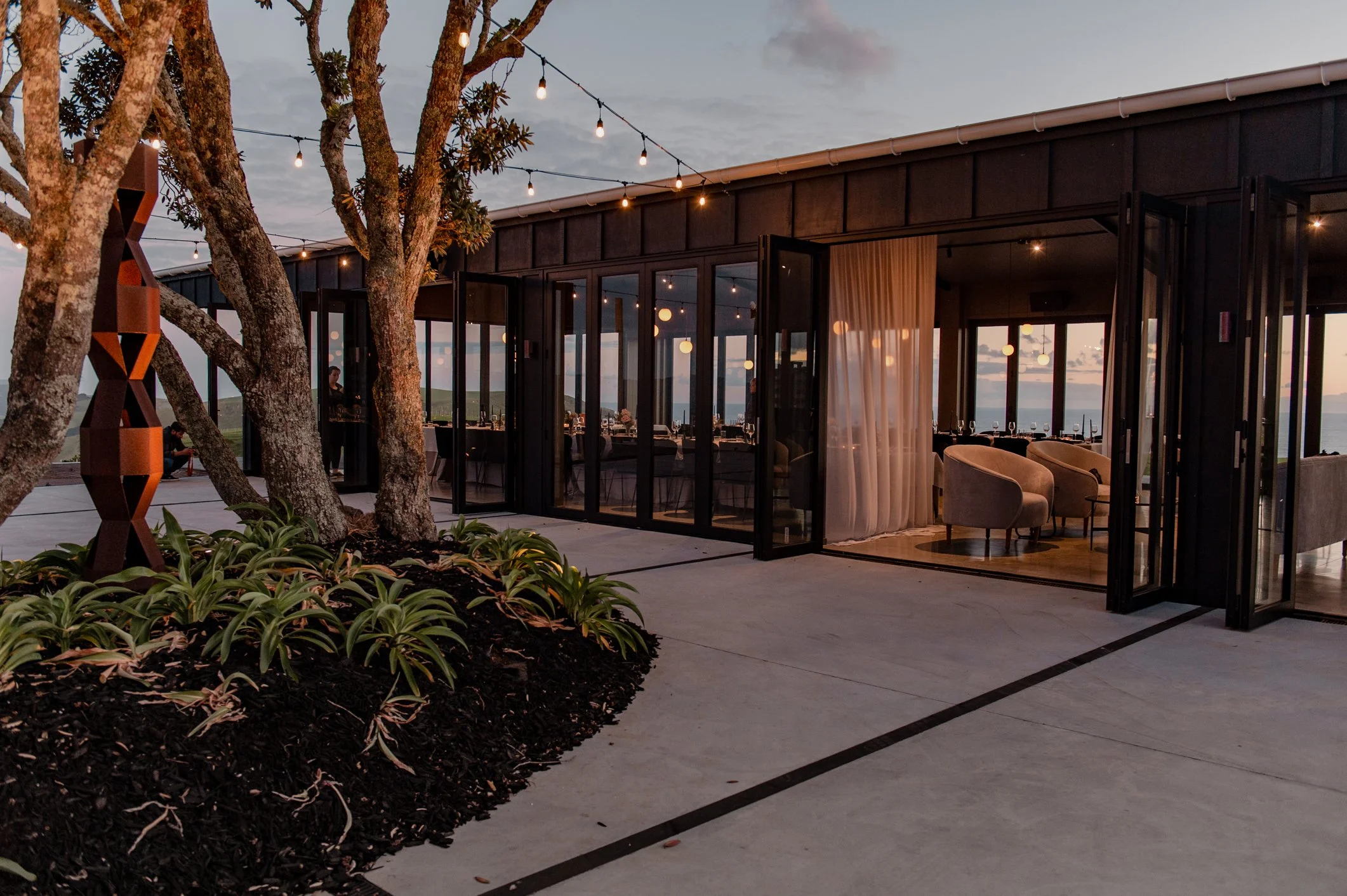 Contemporary oceanfront restaurant patio at dusk with string lights, glass doors, landscaped trees, and indoor dining areas