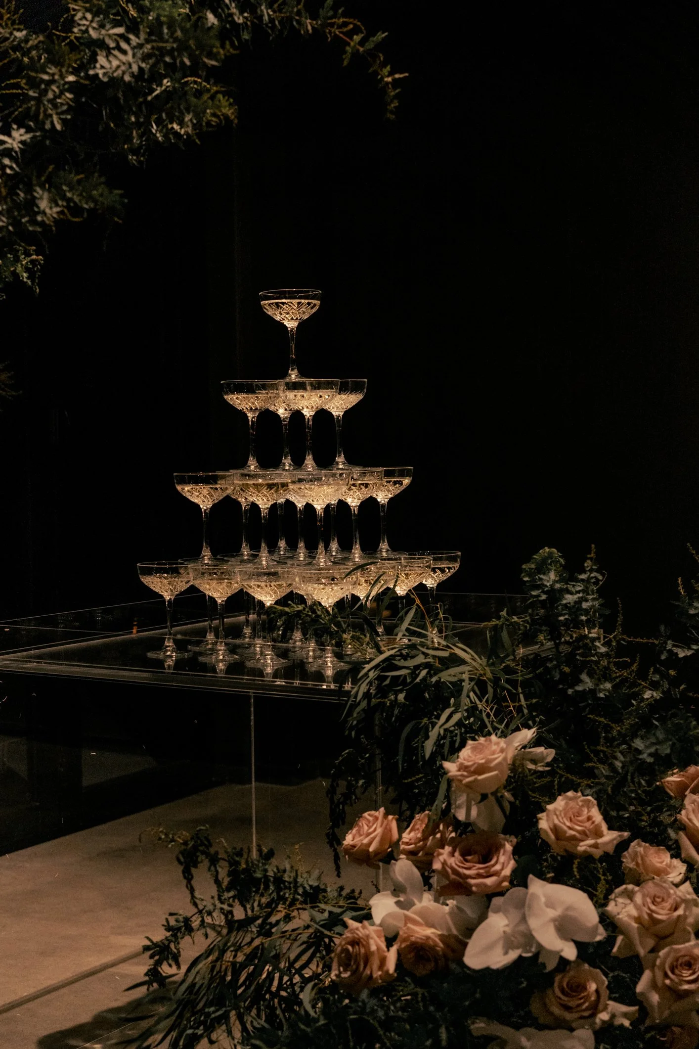 Tiered champagne glass tower surrounded by blush roses and greenery in a dimly lit setting