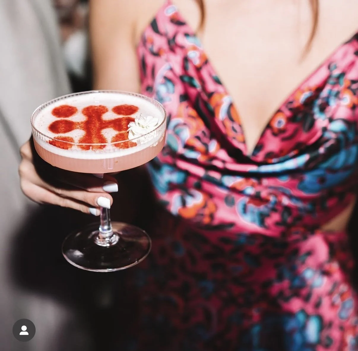 Woman in a colorful floral dress holding a pink cocktail with foam, cinnamon garnish, and a white flower in a coupe glass