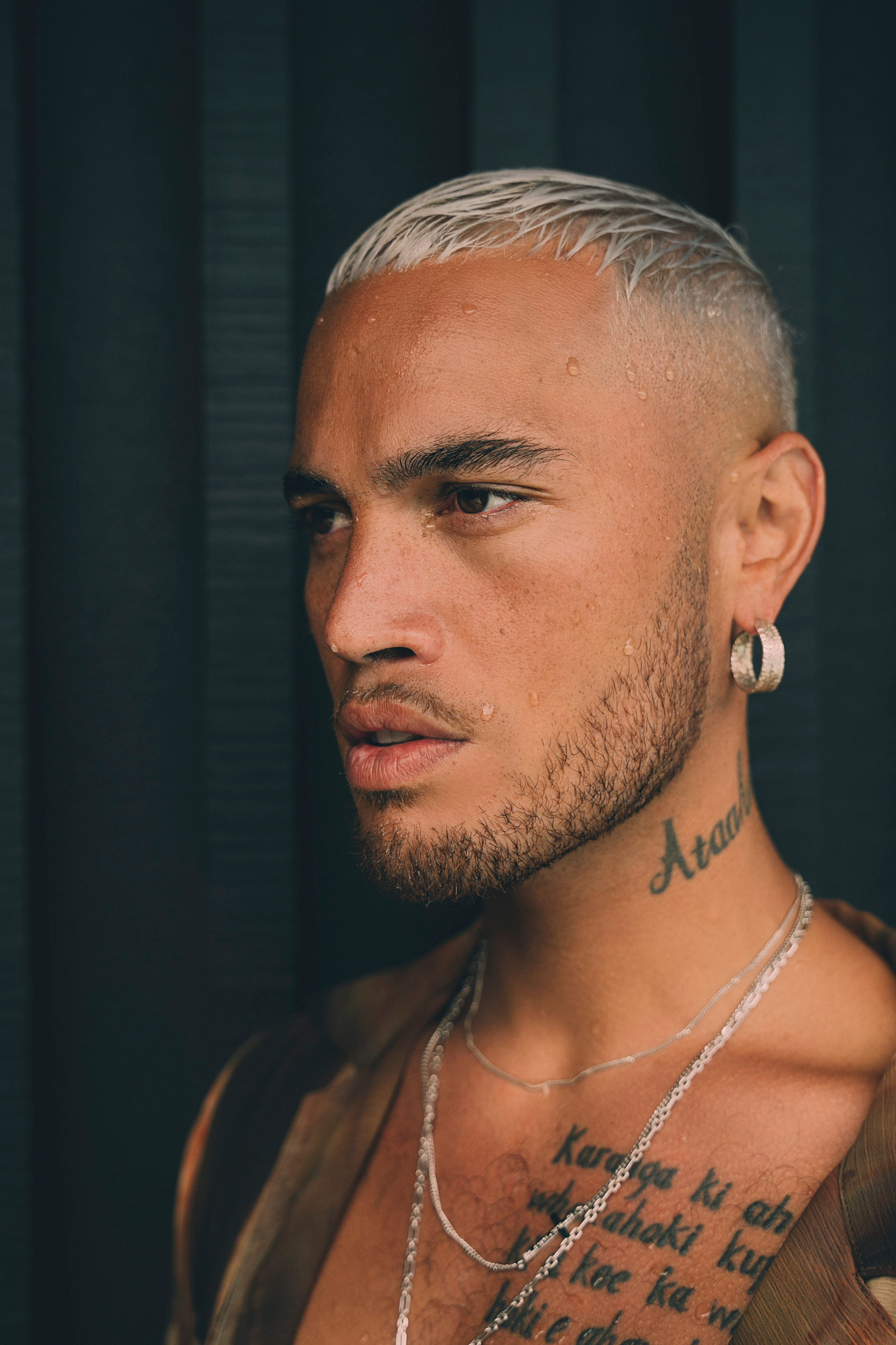 Close-up portrait of a tattooed man with bleached hair, beard, earrings, and layered necklaces