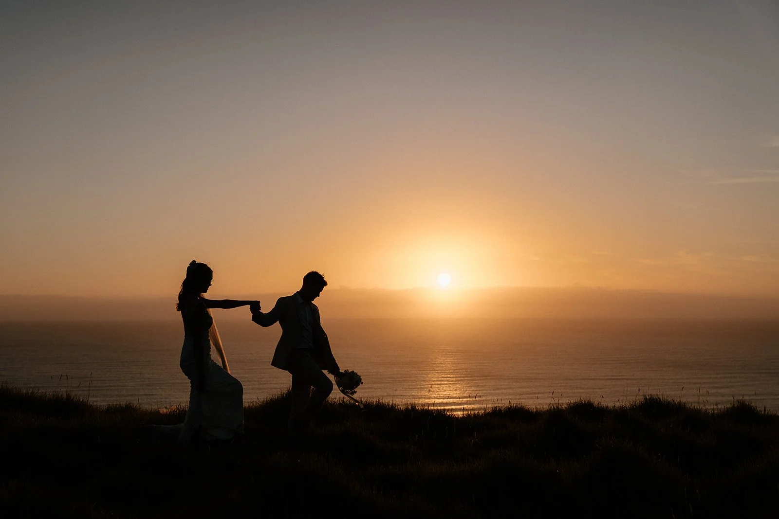 Silhouetted bride and groom holding hands on a grassy seaside cliff at sunset