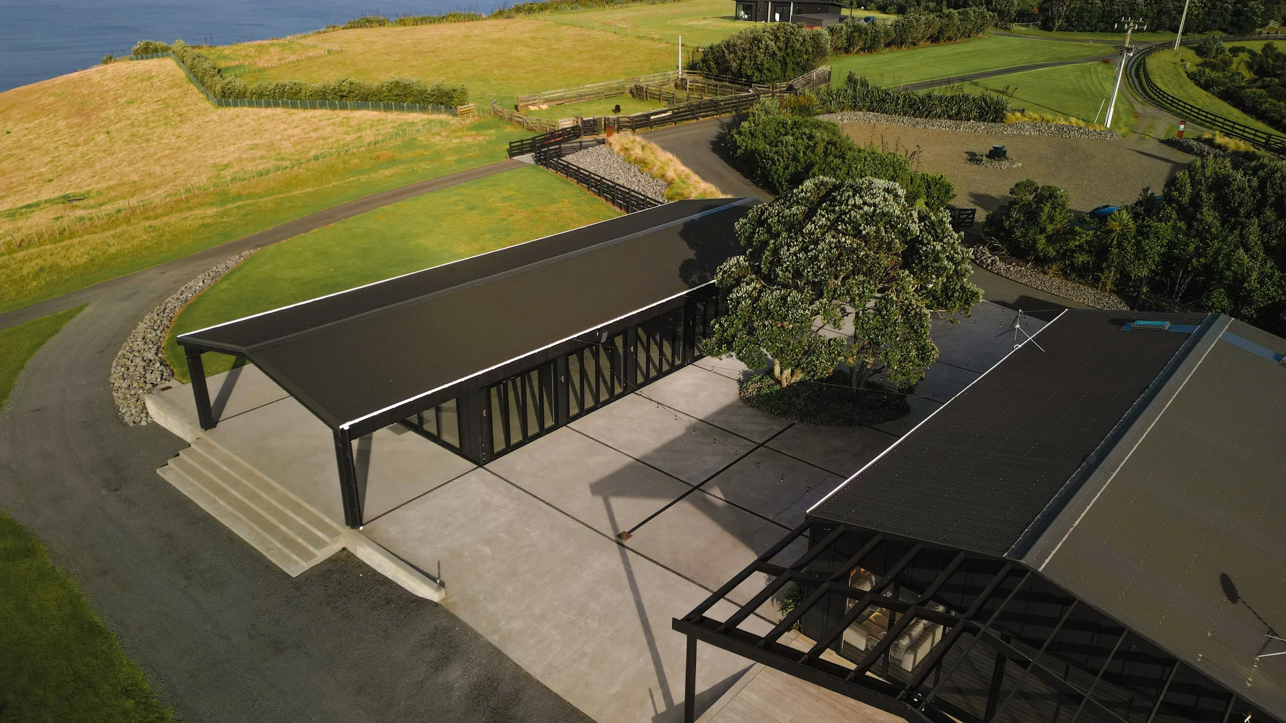 Aerial view of a modern black-roofed property beside a lake, surrounded by manicured lawns, gardens, fences, and rural fields.