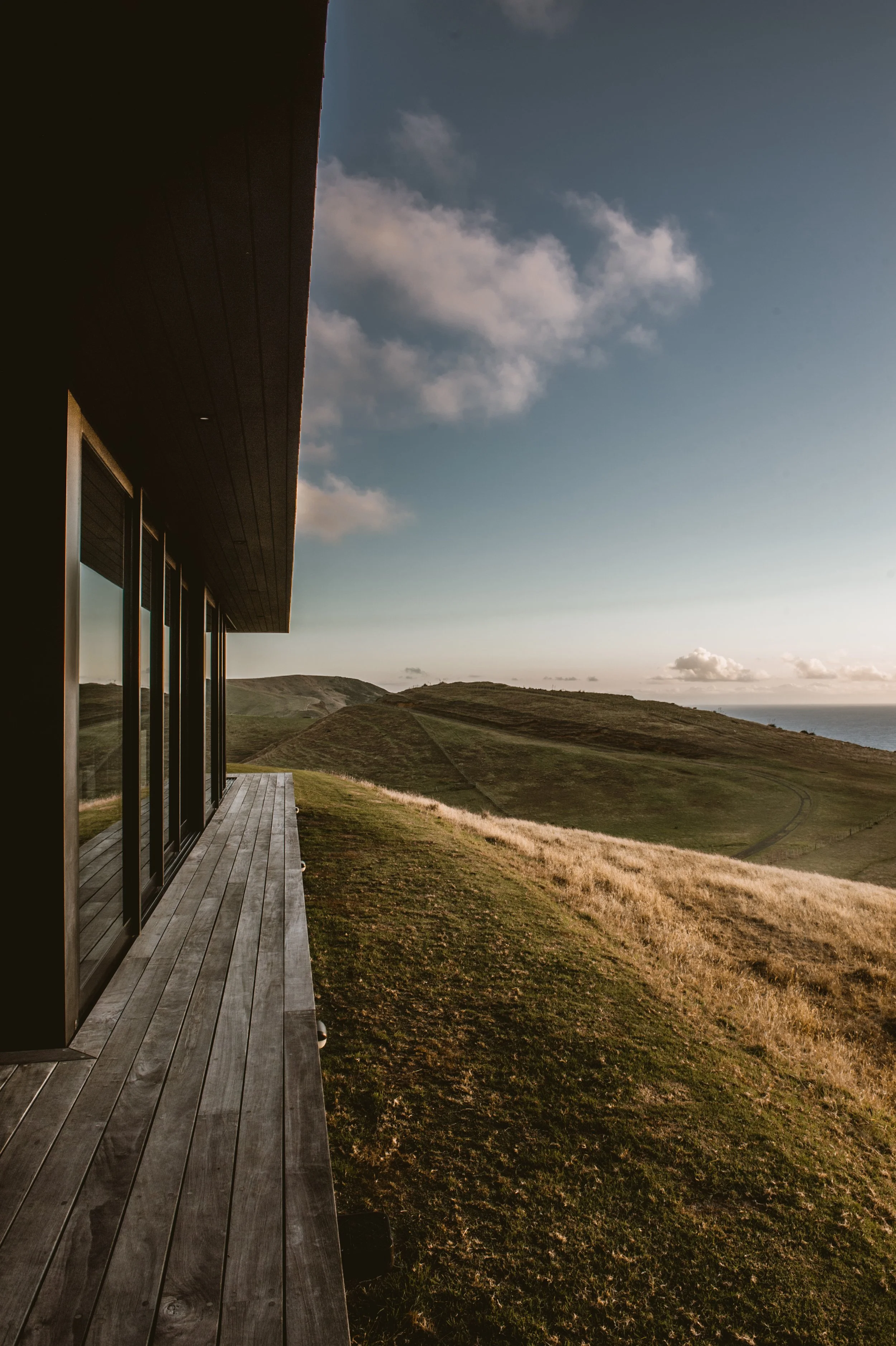 Modern glass-fronted house overlooking grassy coastal hills and the ocean under a cloudy blue sky