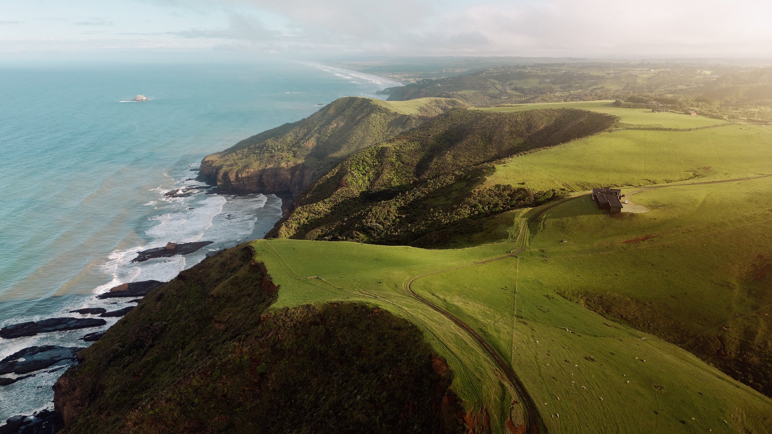 Aerial view of rugged coastal cliffs, turquoise ocean, green farmland, and a small building beneath a hazy sky