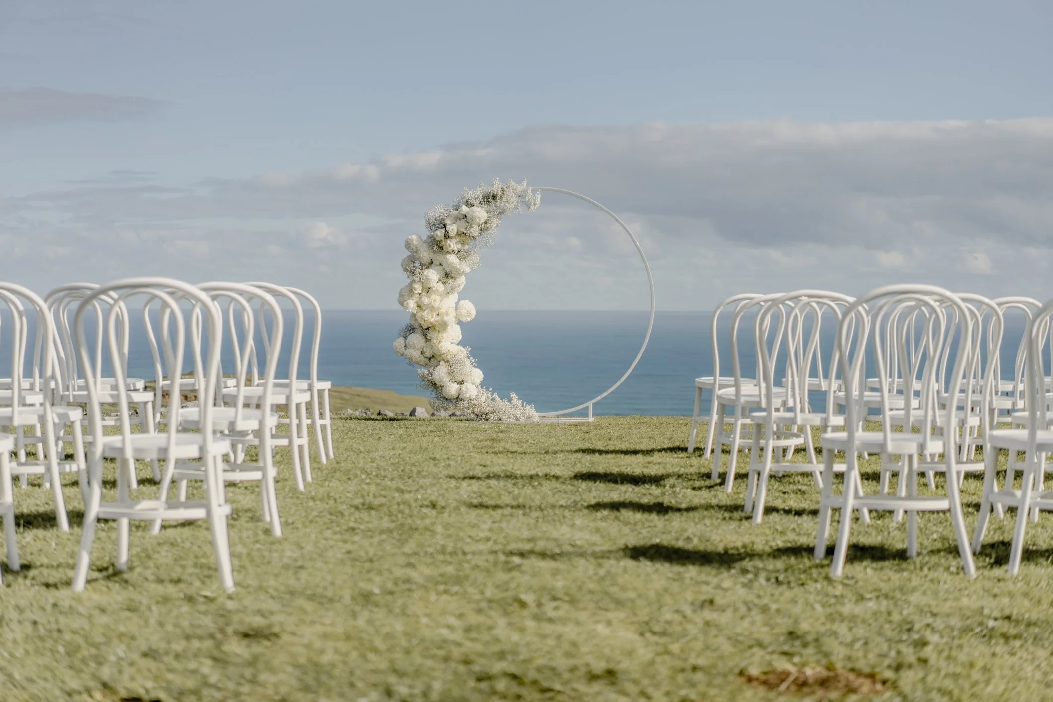 Outdoor wedding ceremony setup with white chairs and a circular floral arch overlooking the ocean