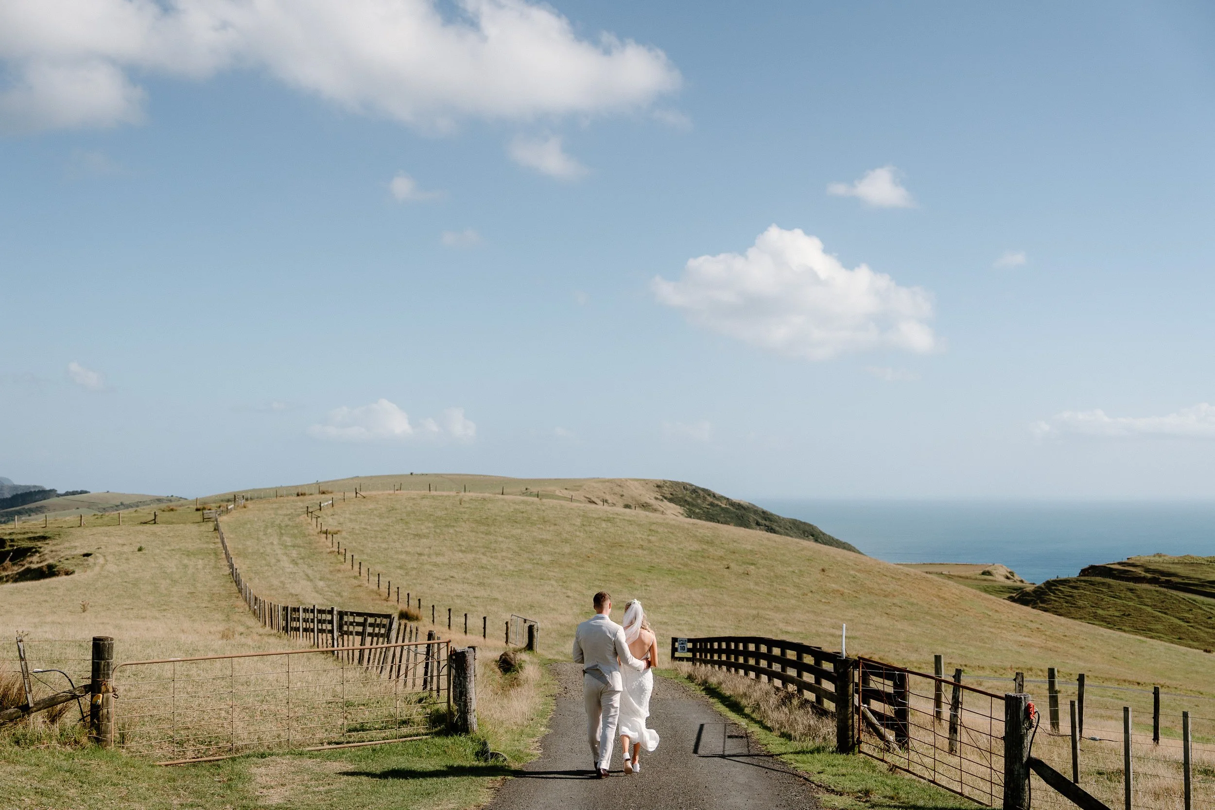 Newlywed couple walking along a fenced coastal countryside path overlooking the ocean