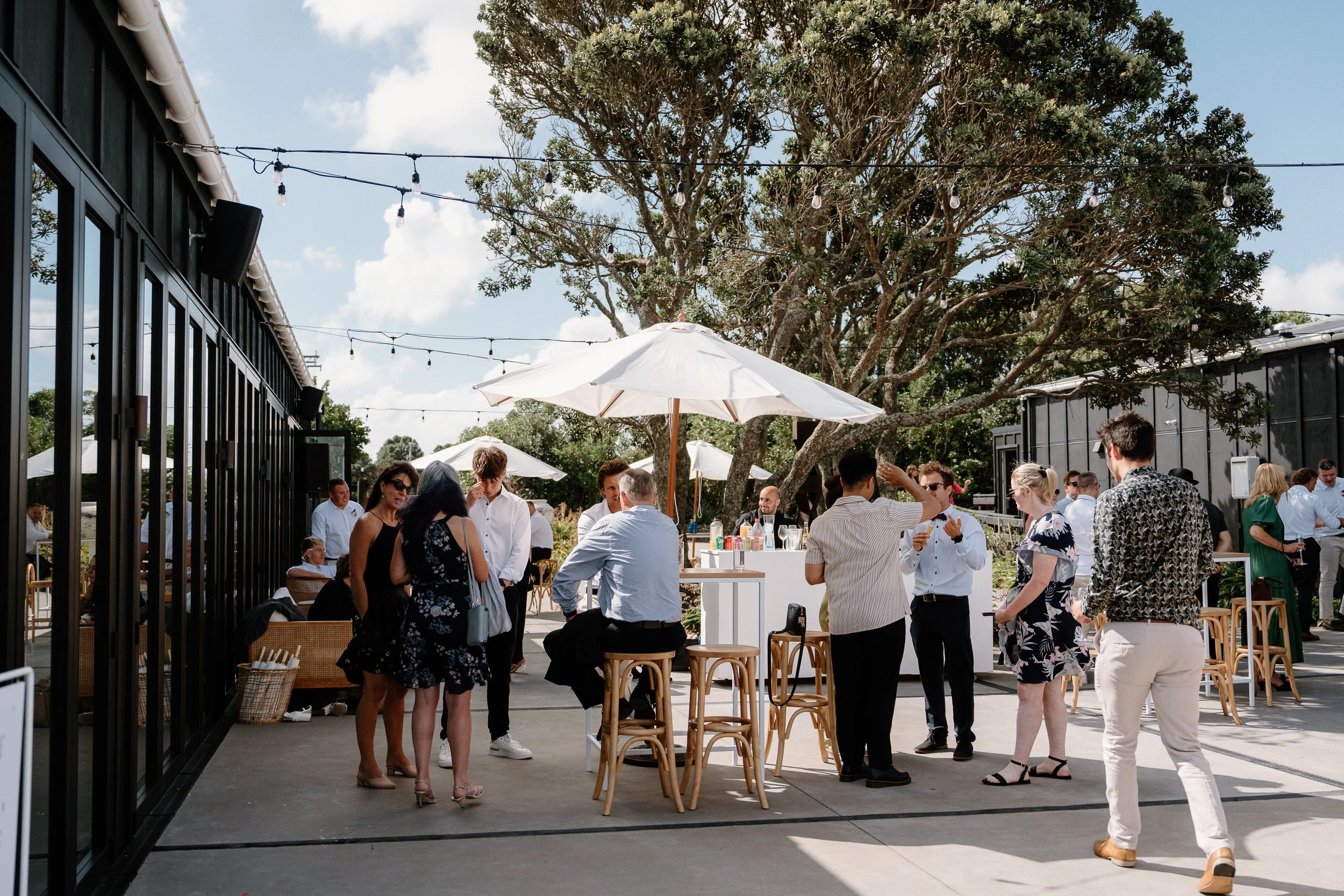 Guests mingling at a sunny outdoor restaurant patio with umbrellas, string lights, and lush trees