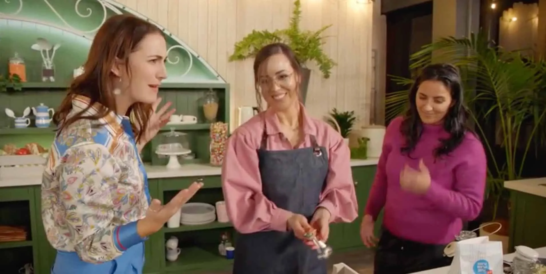 Three women chatting in a warmly decorated kitchen, with one wearing an apron and holding a utensil.
