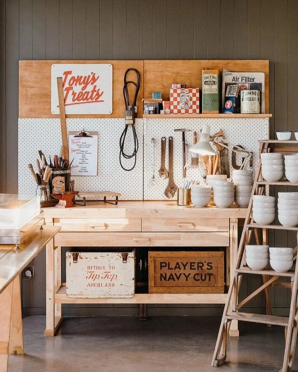 Vintage-style bakery or kitchen workspace with a wooden workbench, pegboard holding utensils, stacked white bowls, baking tools, and retro signs.