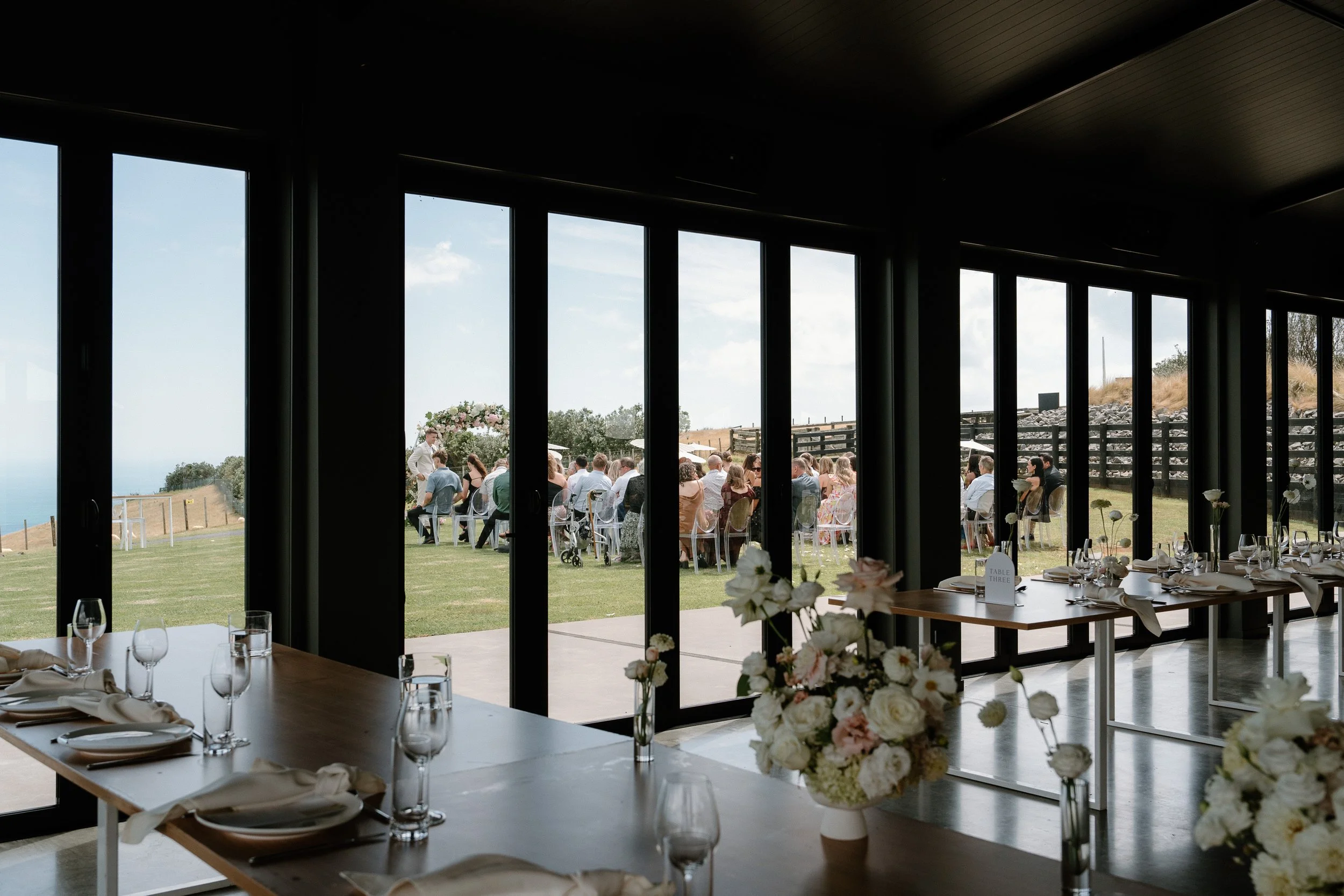 Wedding reception dining room with floral centerpieces and table settings overlooking a coastal outdoor ceremony