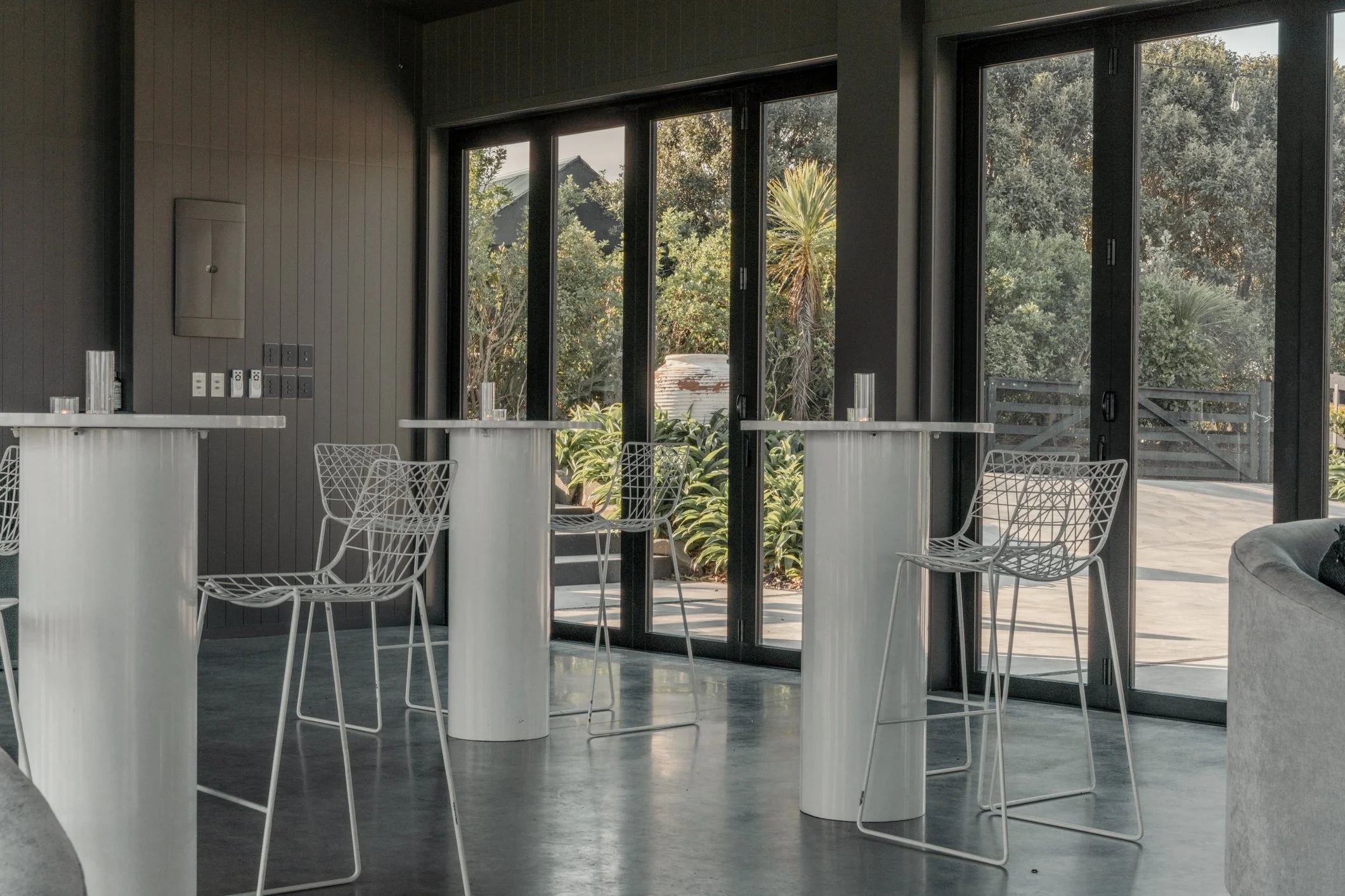 Modern lounge with white wire chairs, cylindrical tables, and floor-to-ceiling glass doors overlooking a landscaped patio