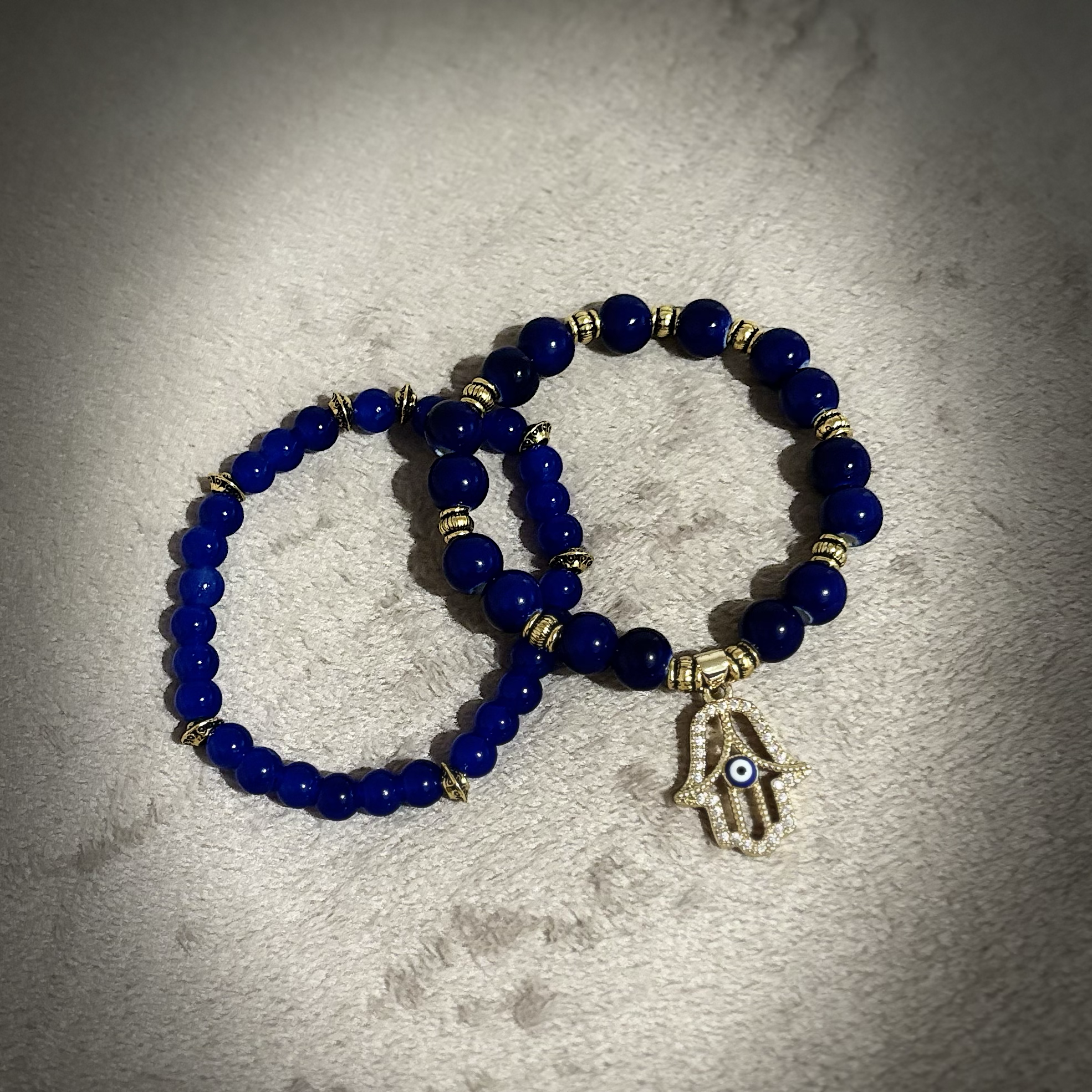Blue beaded bracelet with gold-colored spacers and a gold-colored Hamsa hand charm with an evil eye design, resting on a light-colored surface.