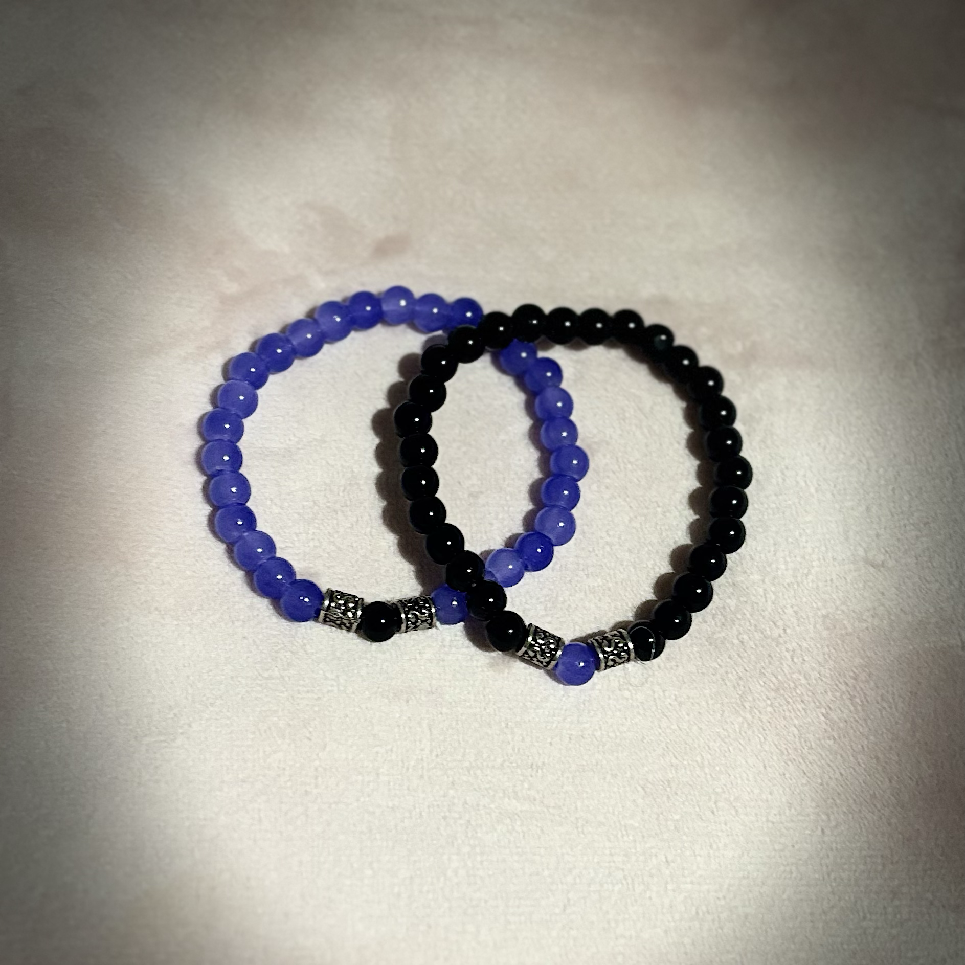 Two beaded bracelets, one with black beads and silver decorative beads, the other with blue beads, on a white textured surface.