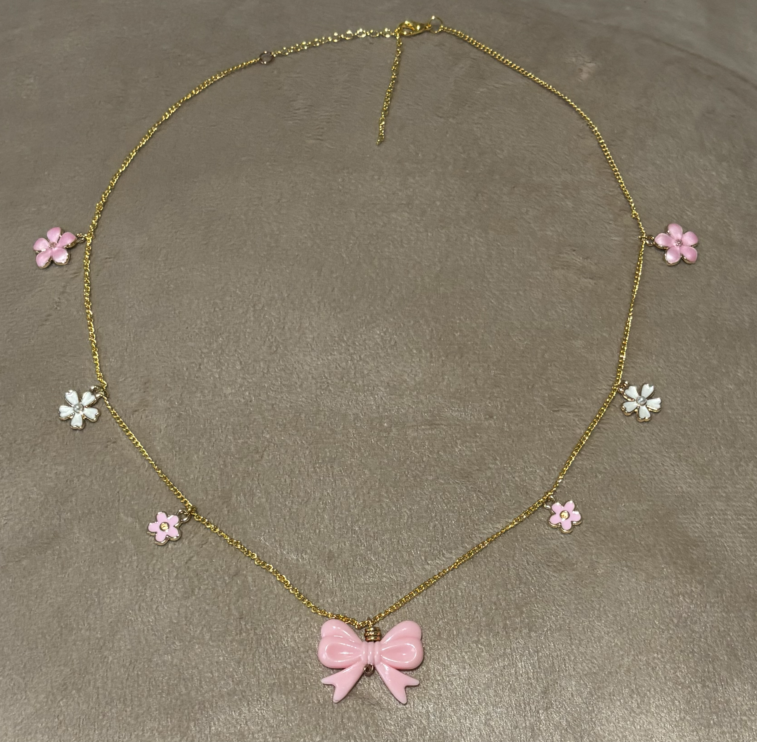 Gold necklace with pink and white flower-themed pendants and a pink butterfly-shaped pendant at the center, laid on a gray surface.