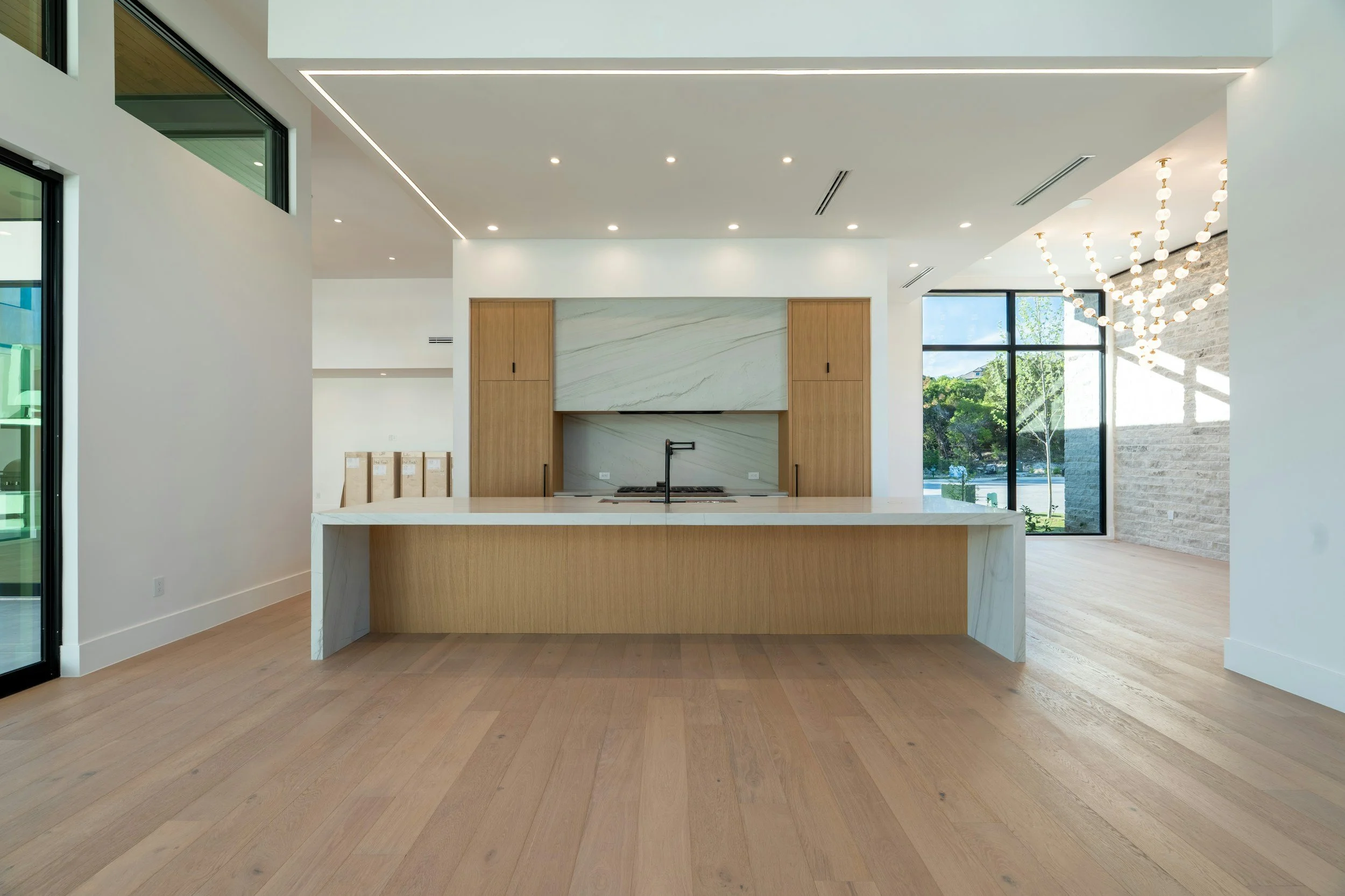 Modern open-concept kitchen with light wood flooring, white walls, large windows, a marble island, and a minimalist design.