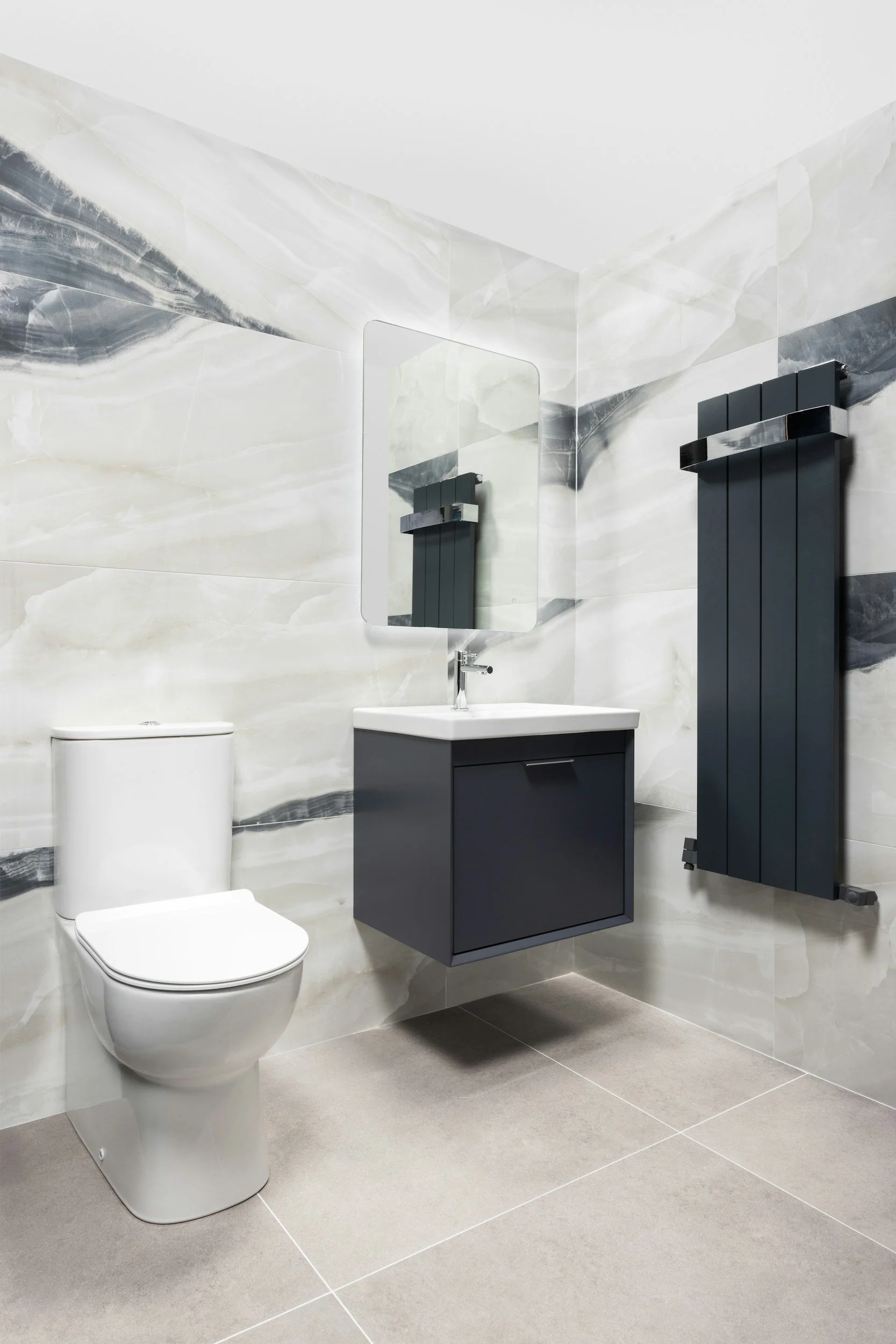 Modern bathroom with a white toilet, a dark gray vanity with a white sink, a rectangular mirror, a wall-mounted black radiator, and marble-like wall tiles.