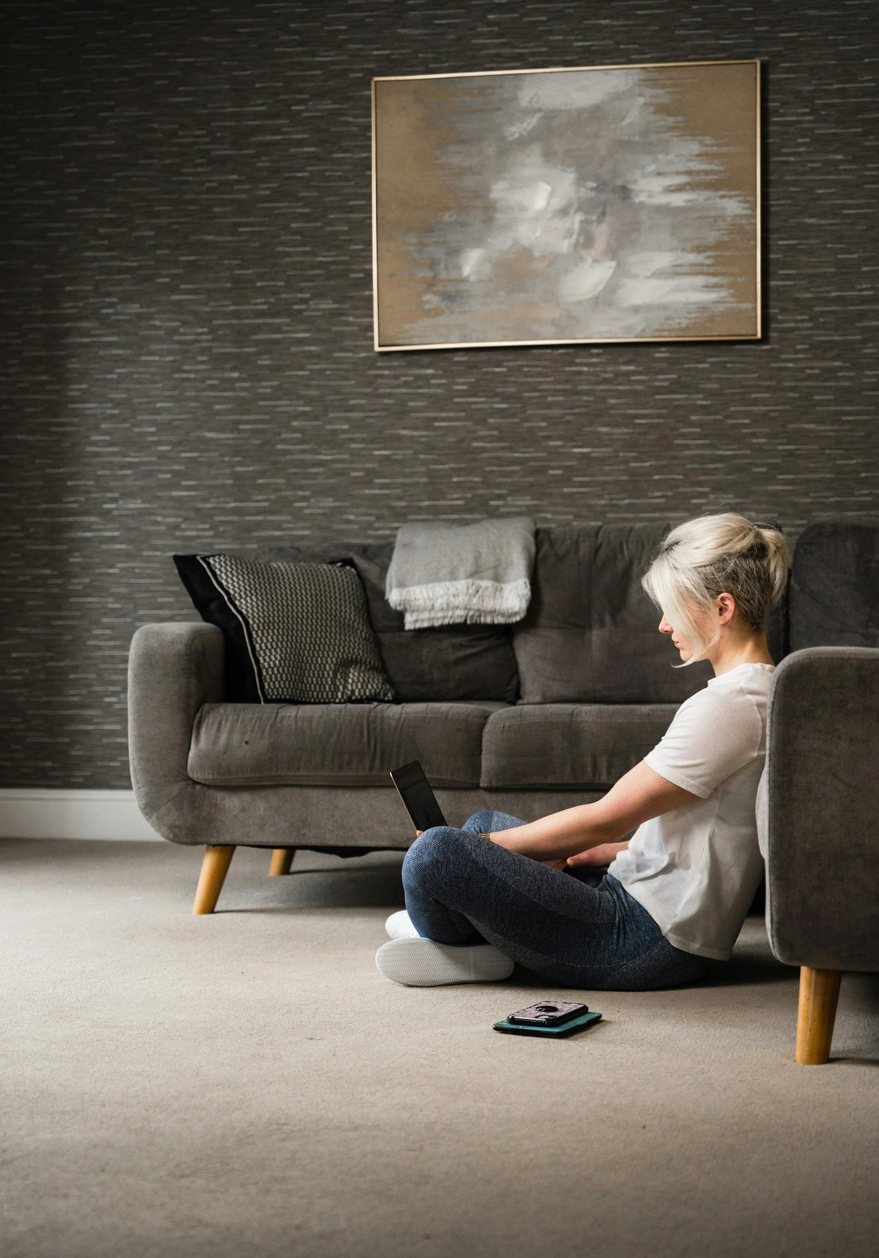 A woman sitting on the floor of a living room, using a smartphone with a child-sized cushion beneath her. There are two stacked phones on the beige carpet, a gray sofa with black and white pillows, a folded blanket, and abstract artwork on the dark textured wall in the background.