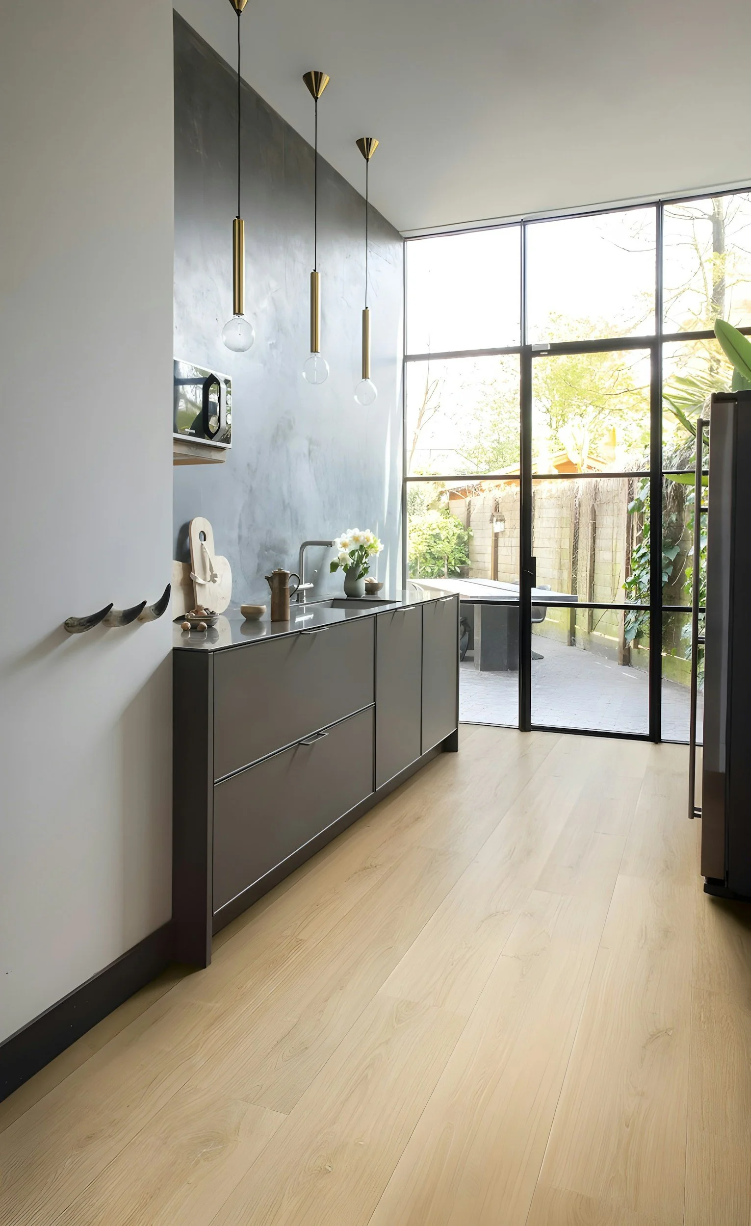 Modern kitchen with gray cabinetry, a window with black framing, pendant lights, and light wood flooring, overlooking backyard with trees and a fence.