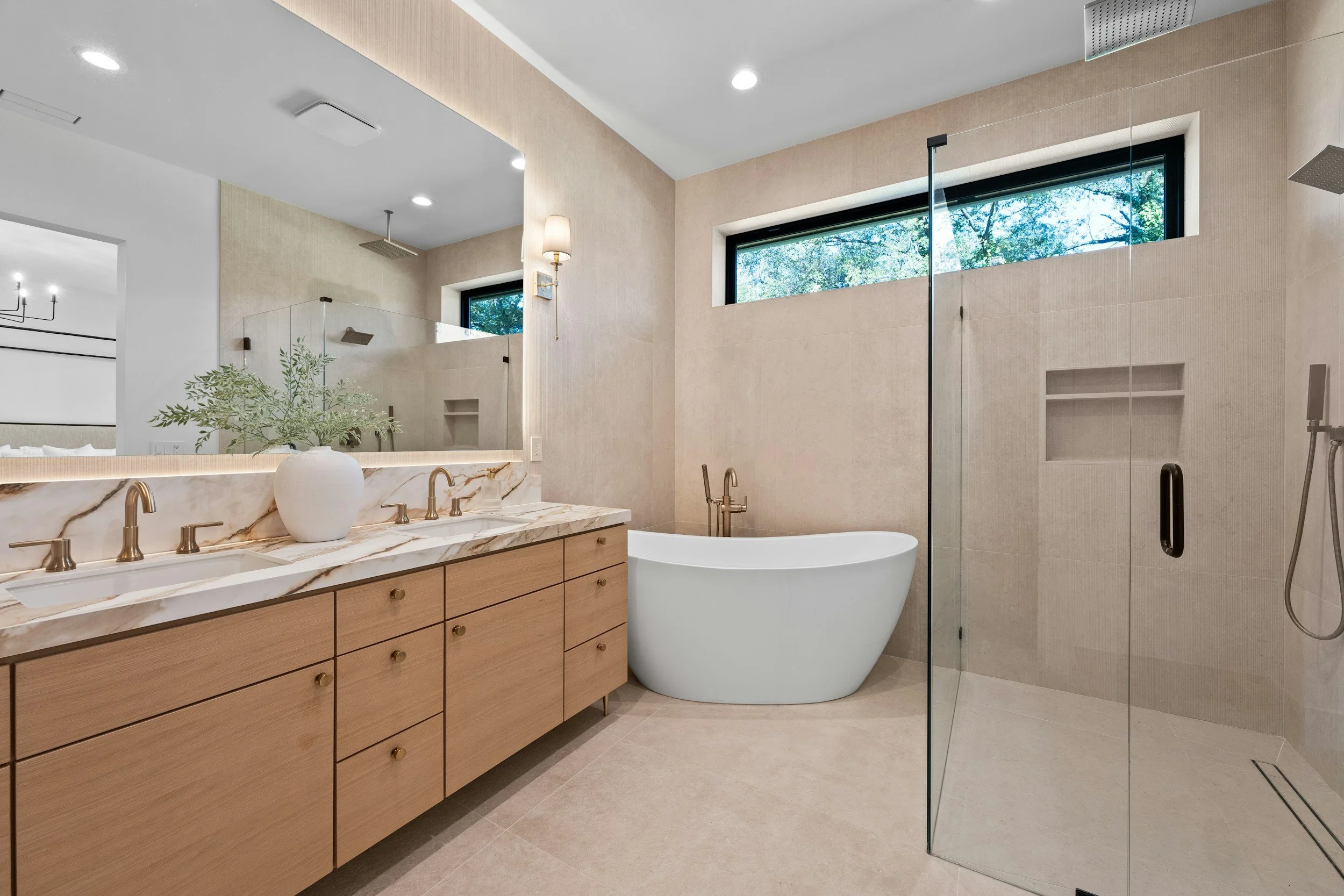 Modern bathroom with beige tiles, a large window showing trees outside, a wooden vanity with a marble countertop, a vessel bathtub, and a glass-enclosed shower area.