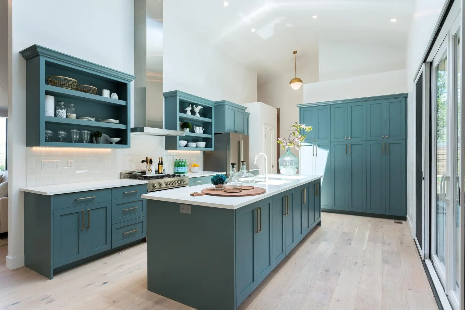 Modern kitchen with blue cabinets, white countertops, open shelving, stainless steel appliances, and a large sliding glass door.