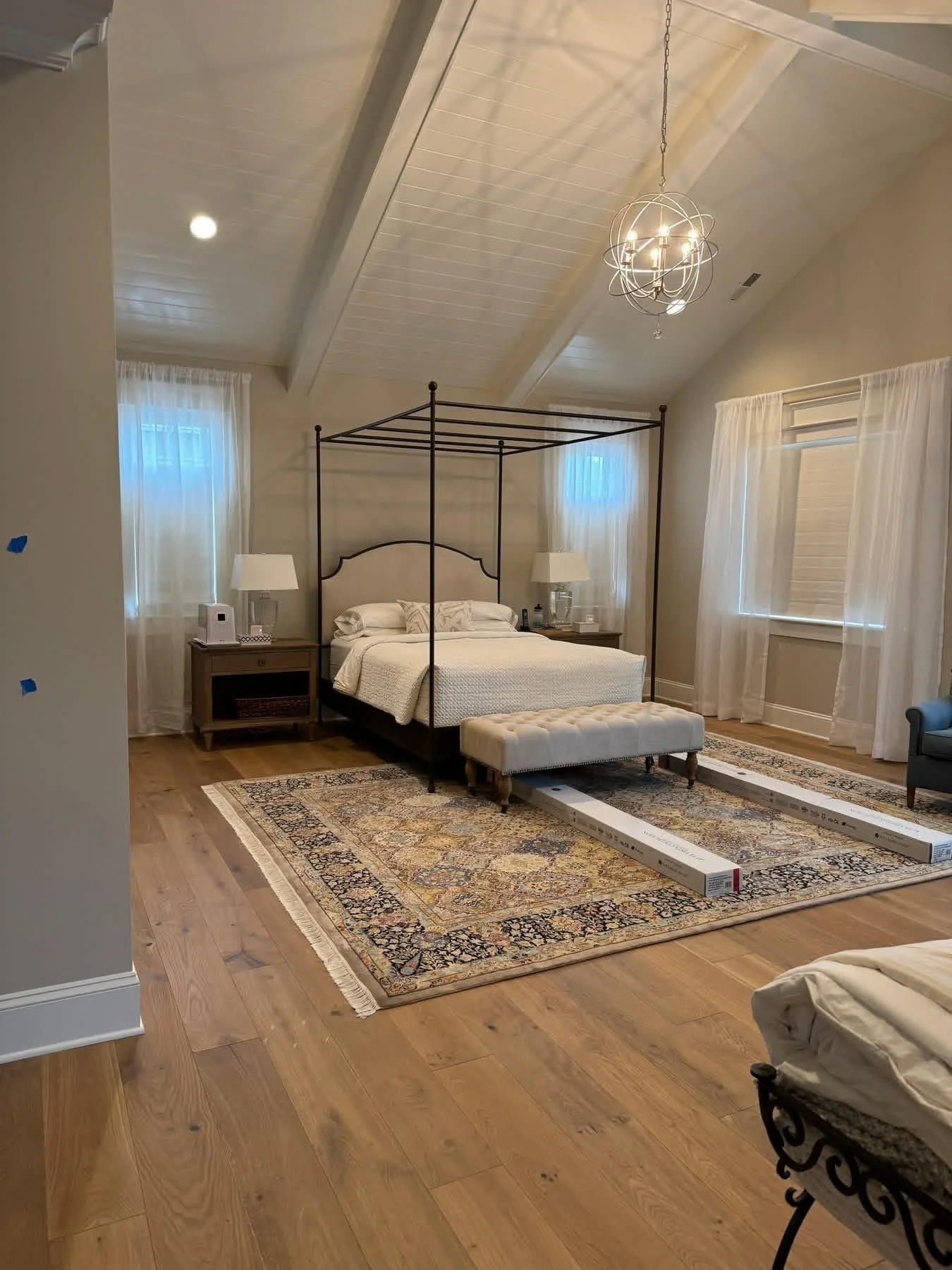 A bedroom with a four-poster bed, two nightstands with lamps, a patterned area rug, and windows with sheer curtains. There is a long boxed item and a tufted bench at the foot of the bed, and a colorful armchair on one side.