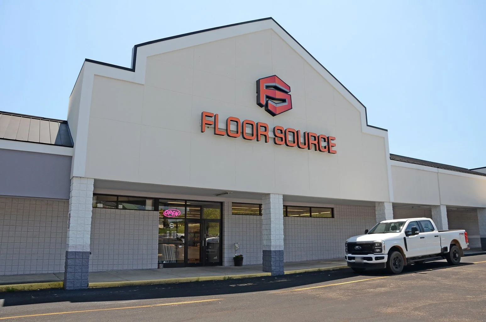 Exterior of a store named Floor Source with a large sign and logo on the building, a white pickup truck parked outside, and a neon 'Open' sign in the window.