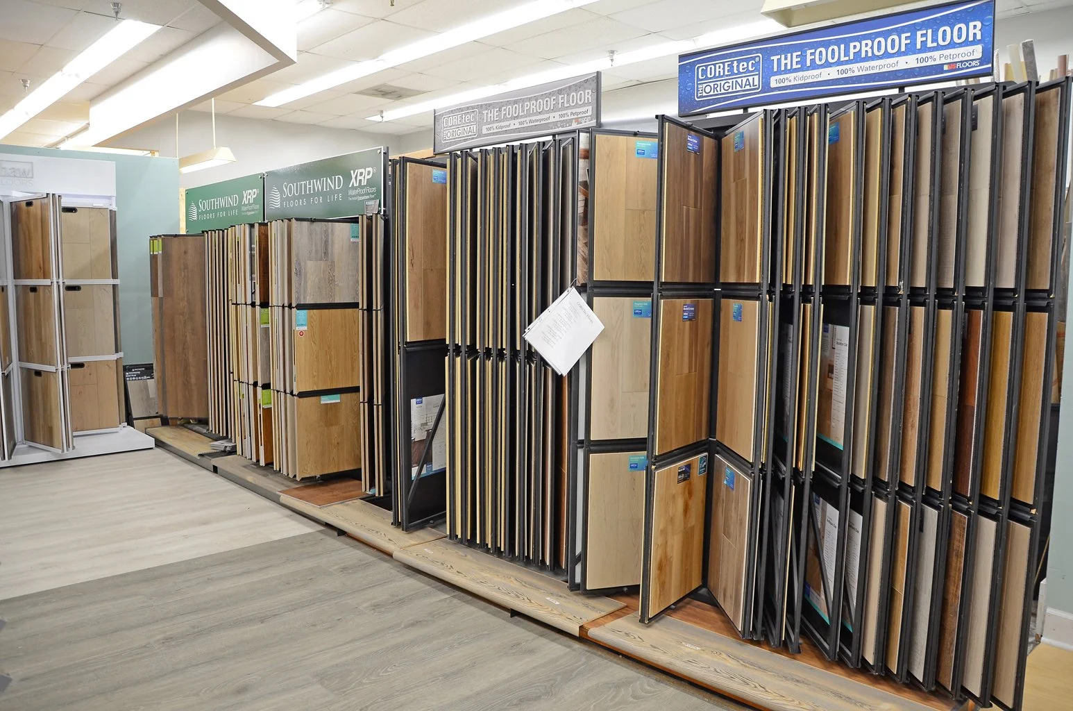 Display of flooring samples in a hardware store, showcasing various wood and laminate flooring options, with signs indicating waterproof and kidproof features.